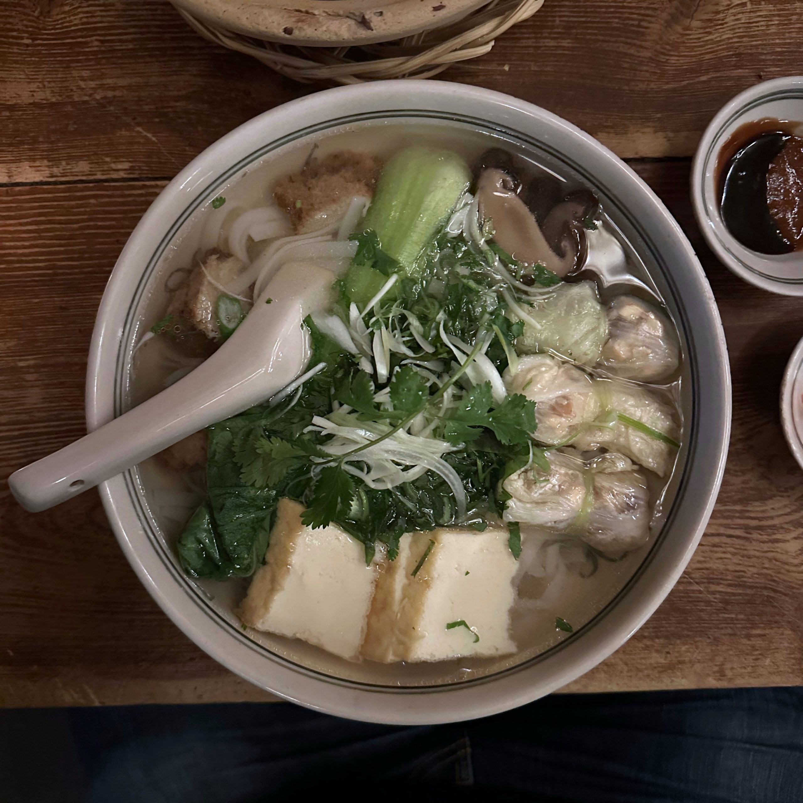 Große Schüssel Pho mit Tofu, Reisbandnudeln, kleinen Kohlrouladen, Shiitake-Pilzen, Pak Choi, Seitanstreifen, frischem Koriander, Frühlingszwiebeln und Sojasprossen in klarer Brühe, serviert mit einem Porzellanlöffel auf einem Holztisch, daneben kleine Schälchen mit Soße.