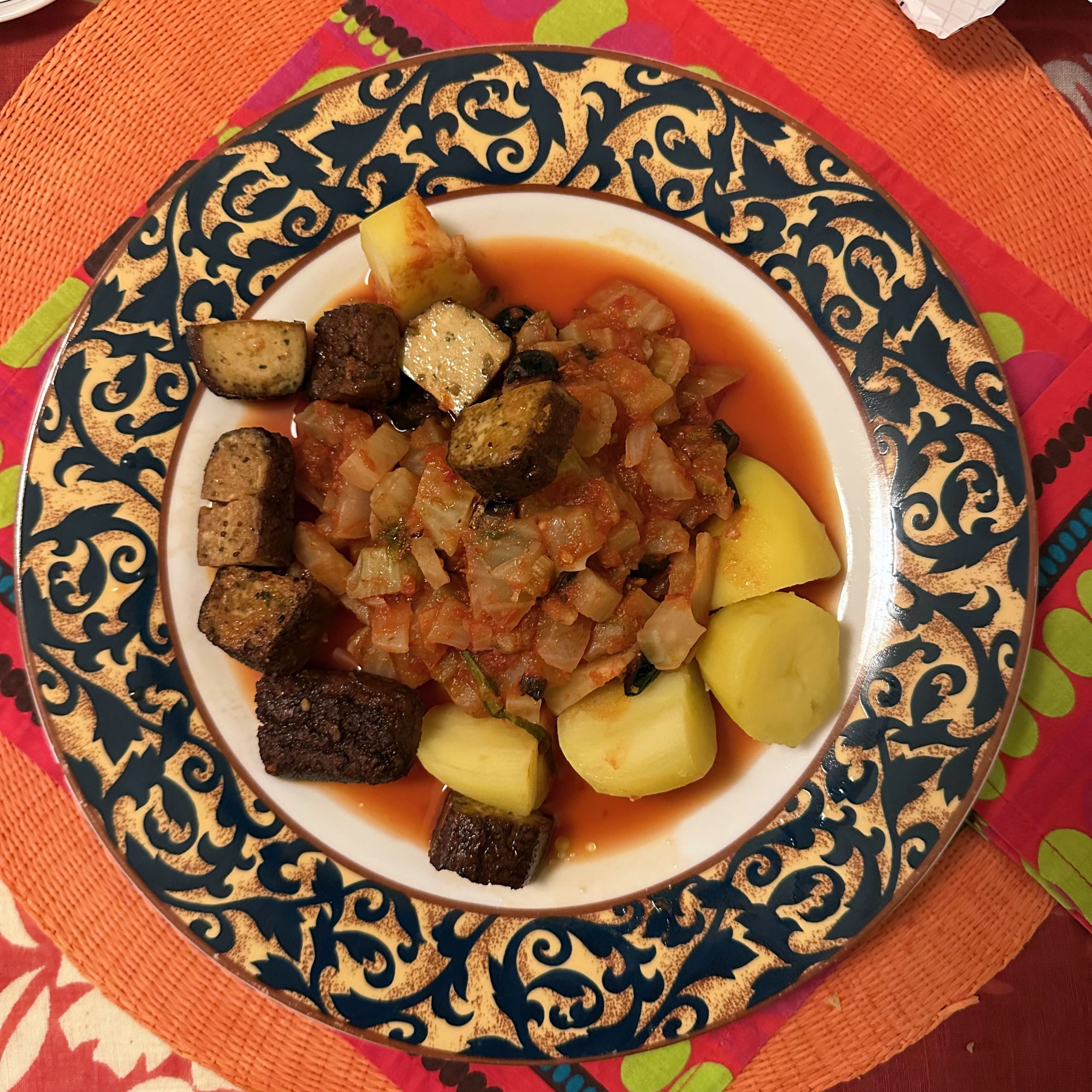 Teller mit Fenchel-Oliven-Tomaten-Ragout, Salzkartoffeln und knusprig gebratenem Räuchertofu auf einem dekorativen blau-goldenen Teller, serviert auf einer bunten Tischdecke.
