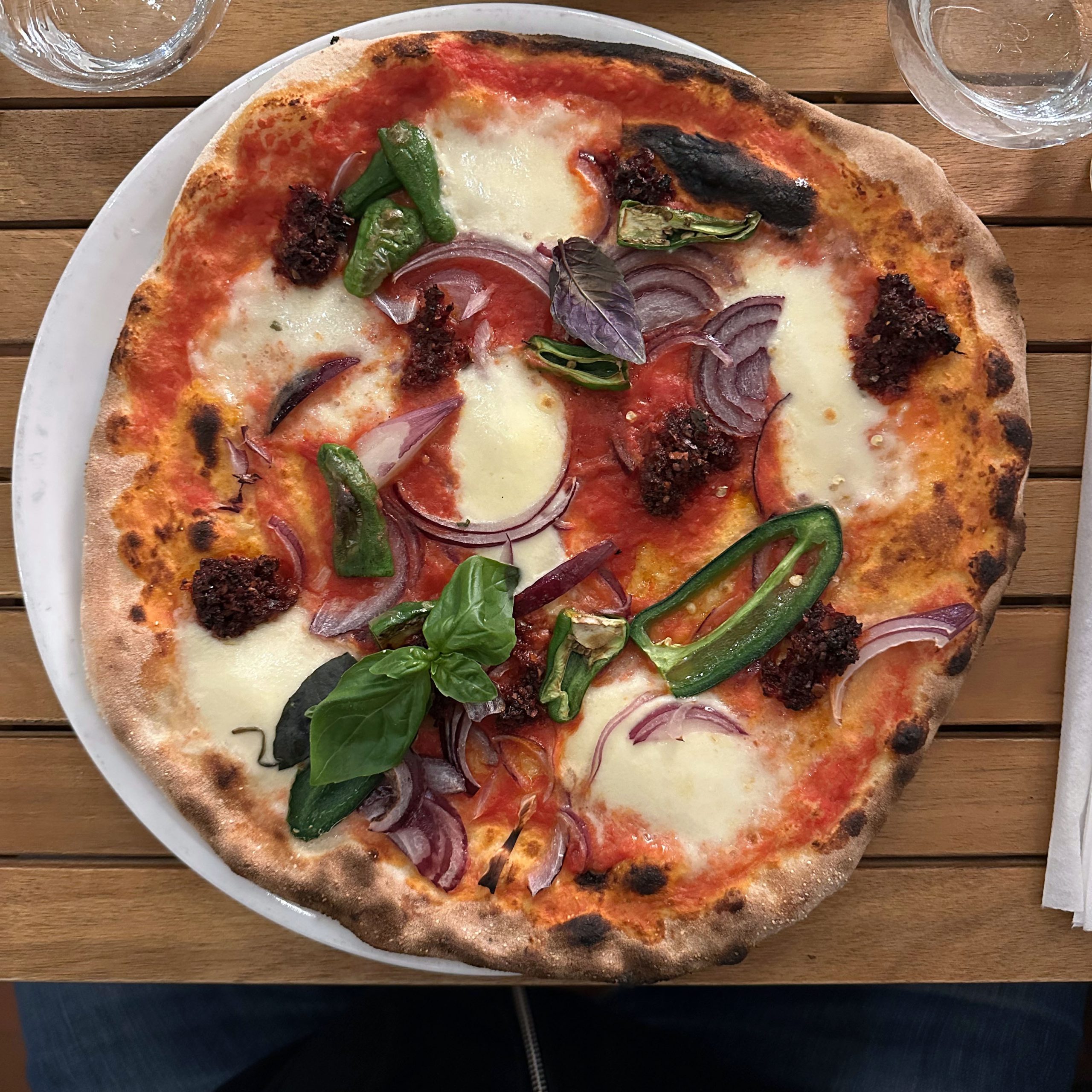 Pizza Bomba im Amo Pizza Romana mit Tomatensauce, Mozzarella, roten Zwiebeln, Nduja, Jalapeños und Basilikum auf einem weißen Teller, serviert auf einem Holztisch.