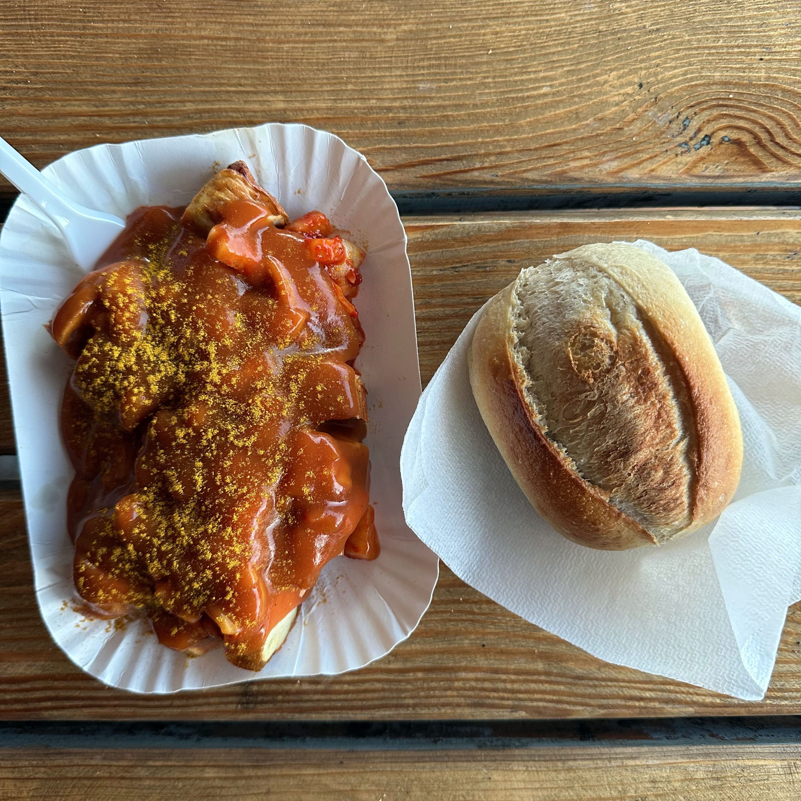 Currywurst mit Currysoße und Currypulver in einer weißen Pappschale, daneben eine Schrippe auf einer Serviette, serviert auf einem Holztisch.