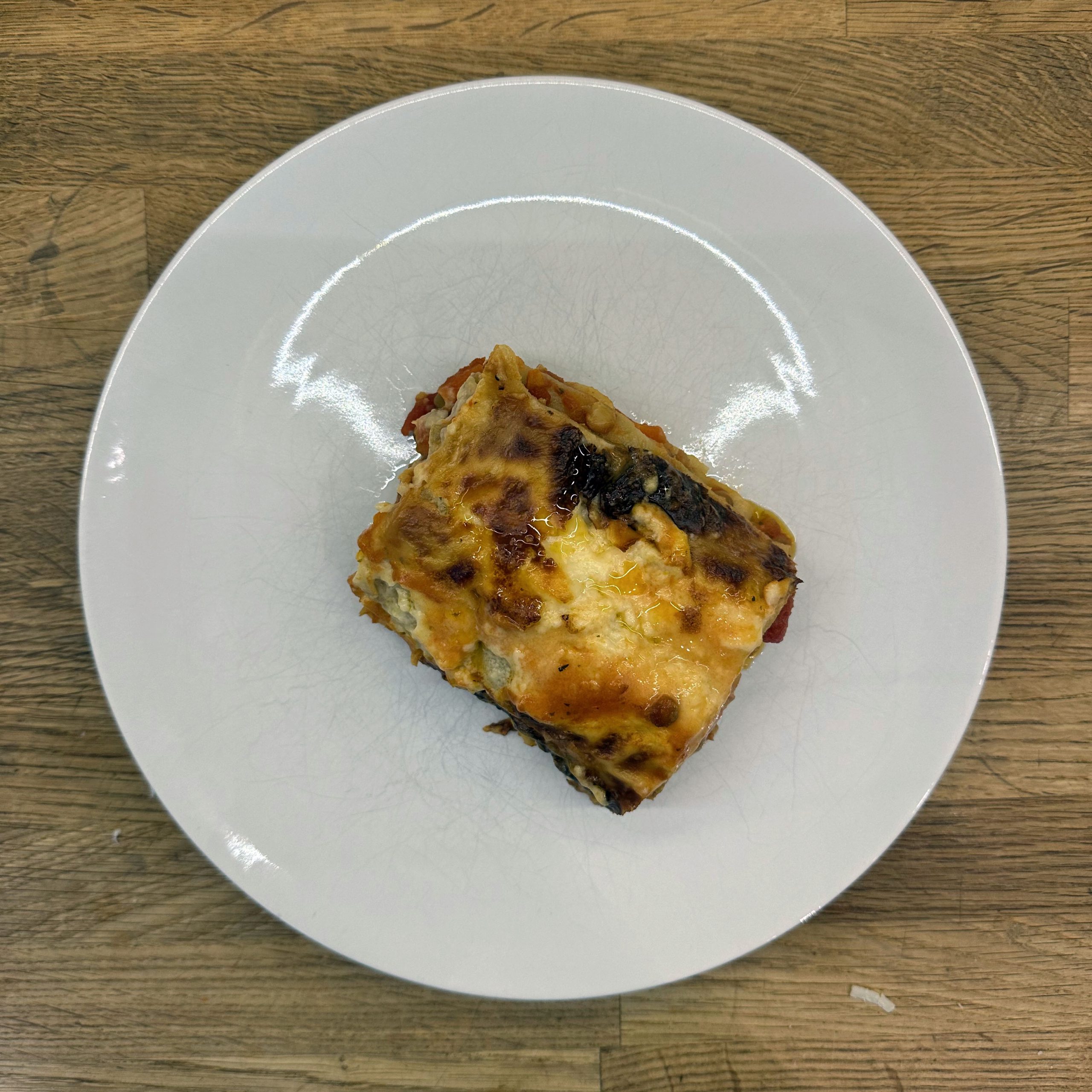 Ein Stück Lasagne mit Linsenbolognese und Béchamelsoße auf einem weißen Teller, von oben fotografiert. Die Oberfläche ist goldbraun gratiniert mit leicht angebräunten Käsestellen. Sichtbare Schichten aus Vollkornlasagneblättern, Linsen-Gemüse-Füllung und heller Béchamelsoße. Der Teller steht auf einem Holztisch.