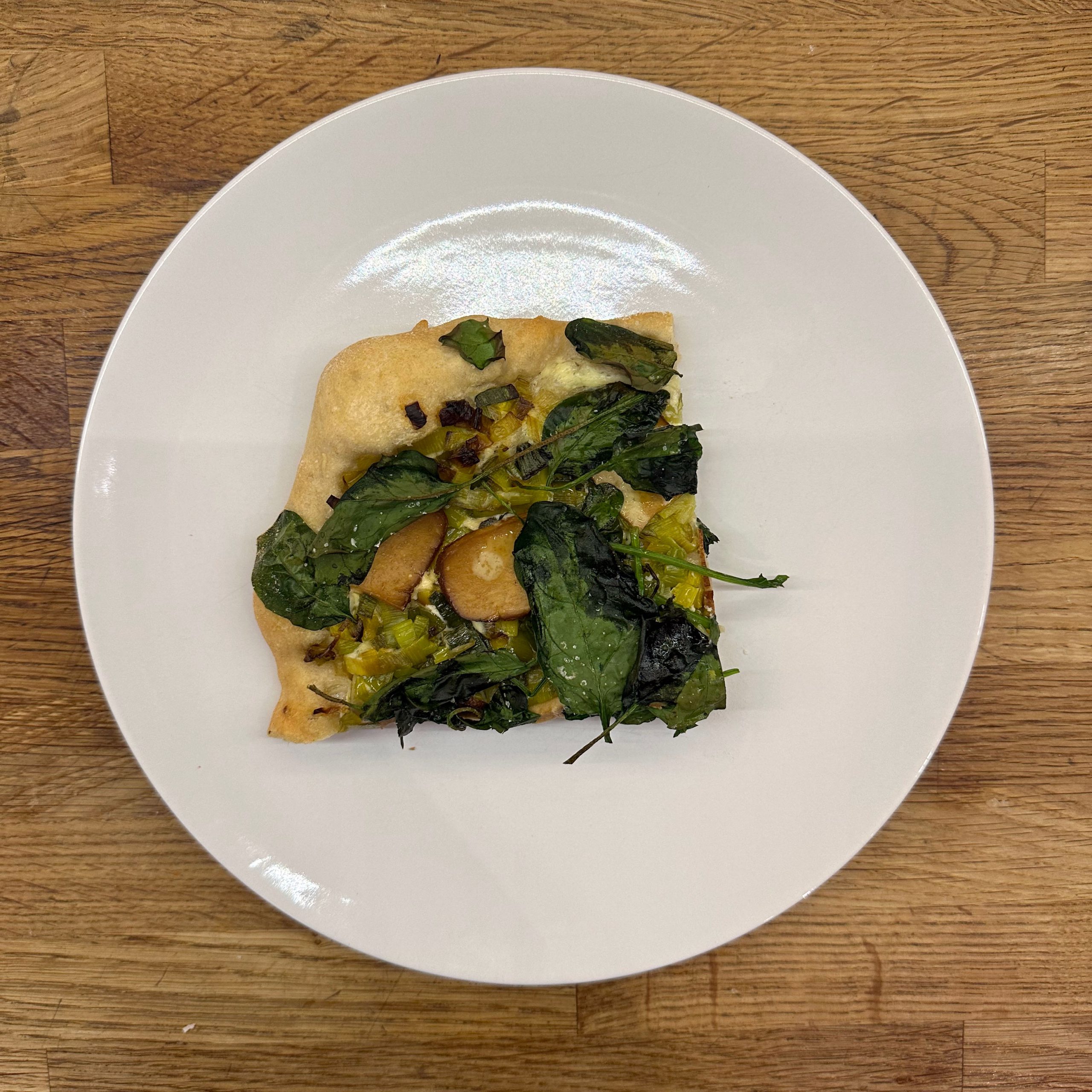 Ein Stück Flammkuchen mit Lauch, Spinat, Feta und Räuchertofu-Scheiben auf einem weißen Teller, von oben fotografiert auf einem Holztisch.