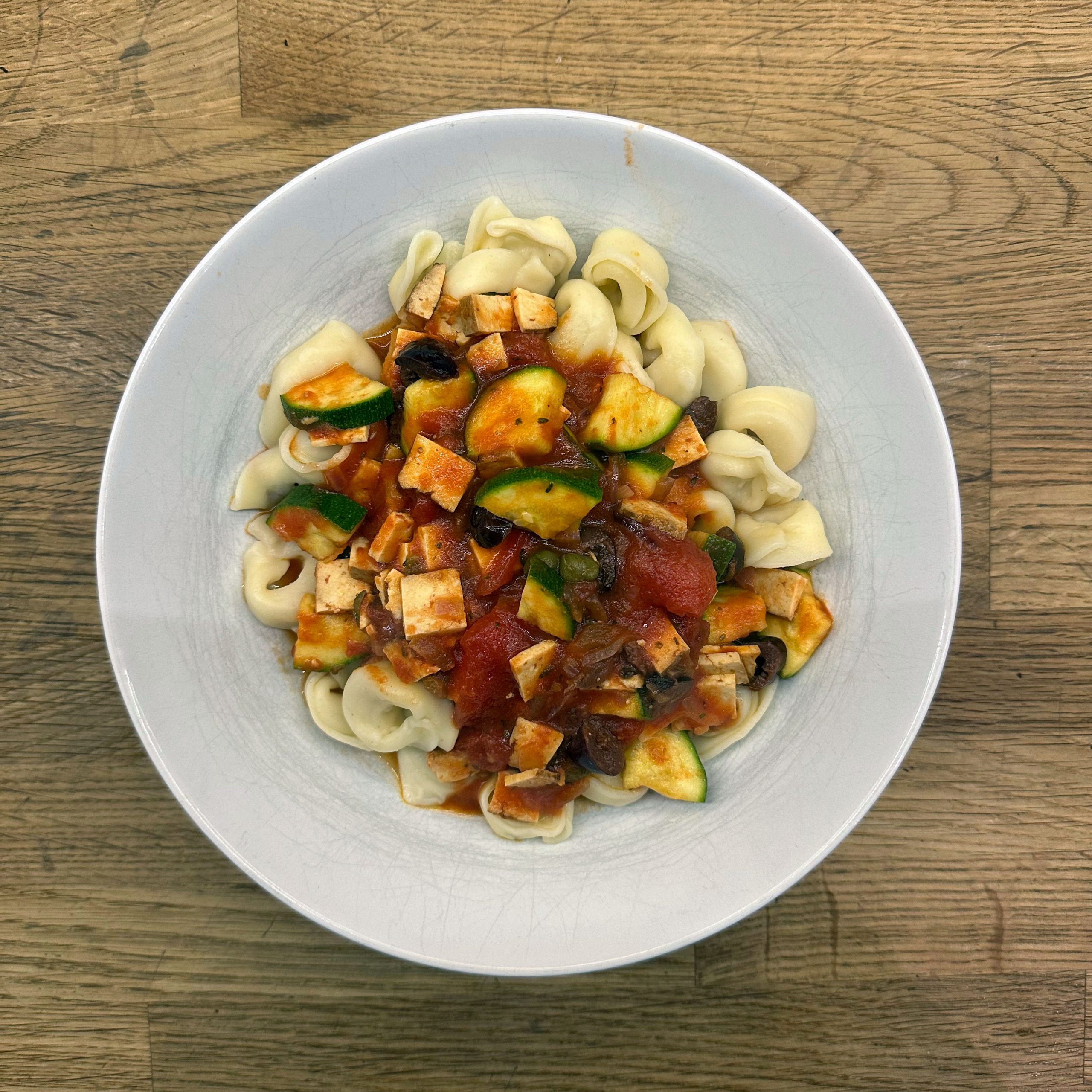 Schüssel mit Gemüsetortellini, übergossen mit einer Tomatensoße mit Räuchertofuwürfeln, Zucchinischeiben, Oliven und Kapern, serviert in einem hellgrauen tiefen Teller auf einem Holztisch.