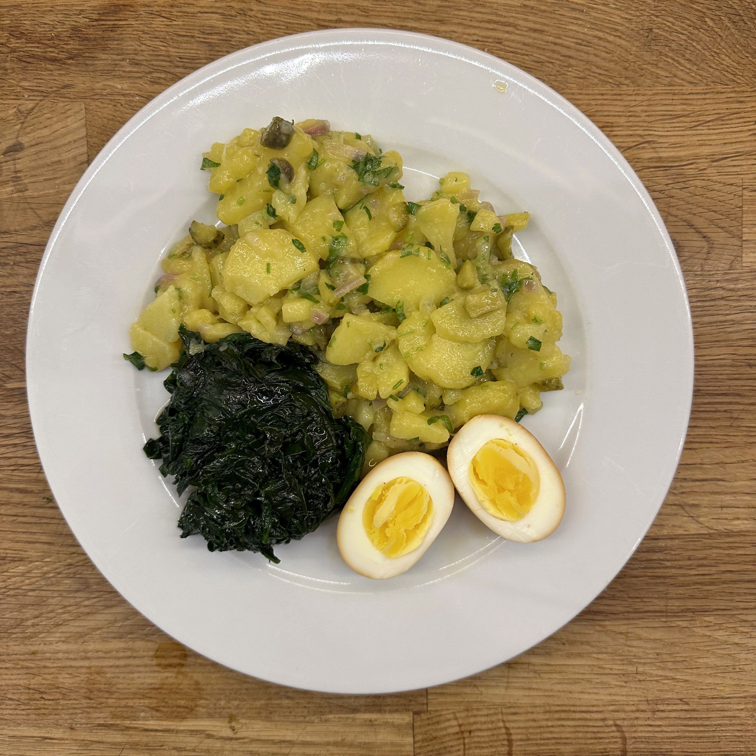 Teller mit schwäbischem Kartoffelsalat mit Petersilie und Zwiebeln, einer Portion gedünstetem Spinat und einem halbierten, in Sojasoße eingelegten Ei auf einem Holztisch.