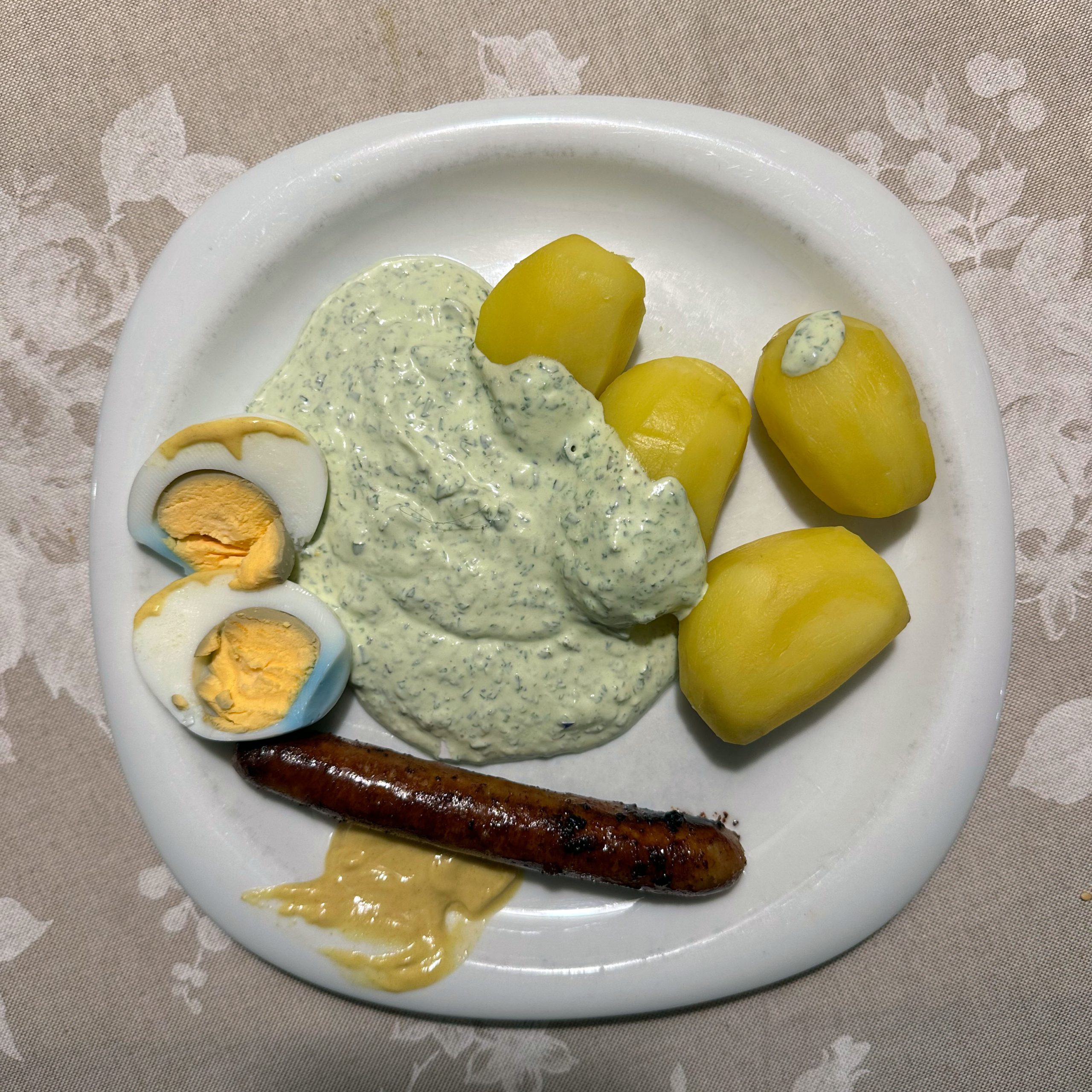 Teller mit Kasseler Grüner Soße, vier geschälten Salzkartoffeln, einem halbierten hartgekochten Ei, einer gebratenen Bratwurst und einem Klecks Senf auf einem weißen Teller.
