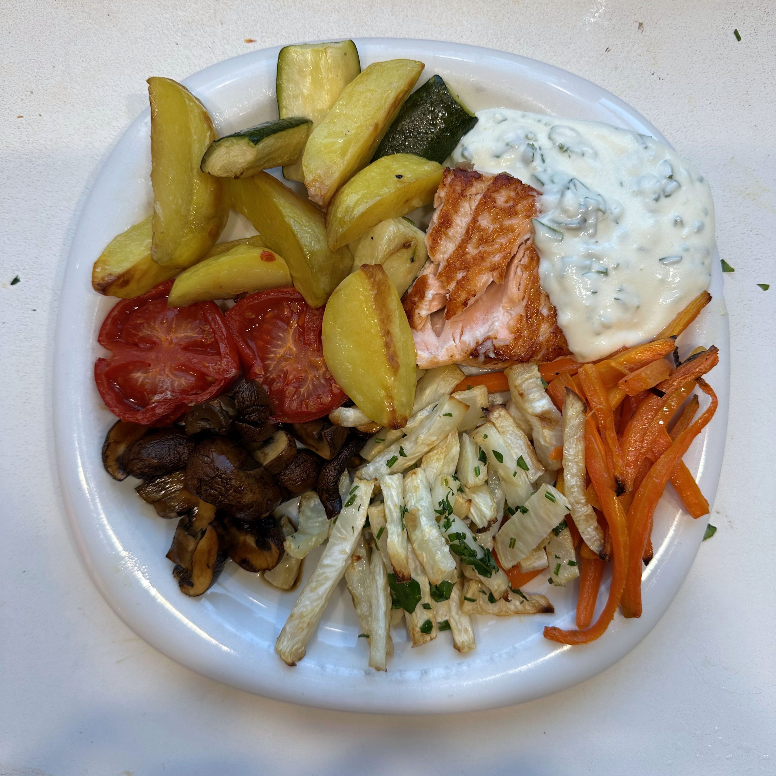 Teller mit gebratenem Lachsfilet, Joghurt-Dip, Ofenkartoffeln, gerösteten Zucchinischeiben, Ofentomaten, marinierten Champignons, Knollenselleriestreifen mit Kräutern und marinierten Paprikastreifen auf weißem Teller.
