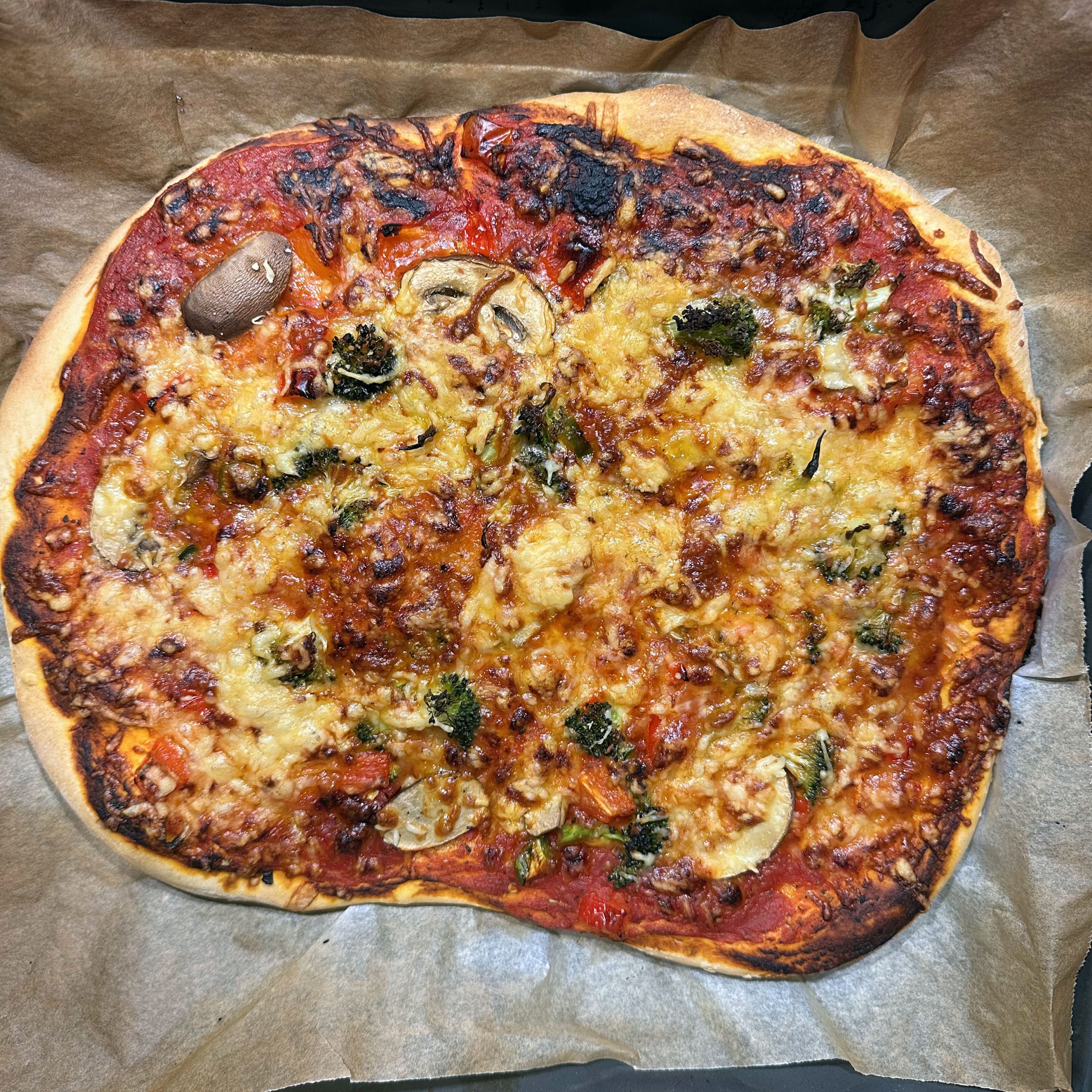 Selbstgemachte Pizza auf Backpapier mit Tomatensoße, geschmolzenem Käse, Champignons, Brokkoli und Paprika, an den Rändern leicht verbrannt.