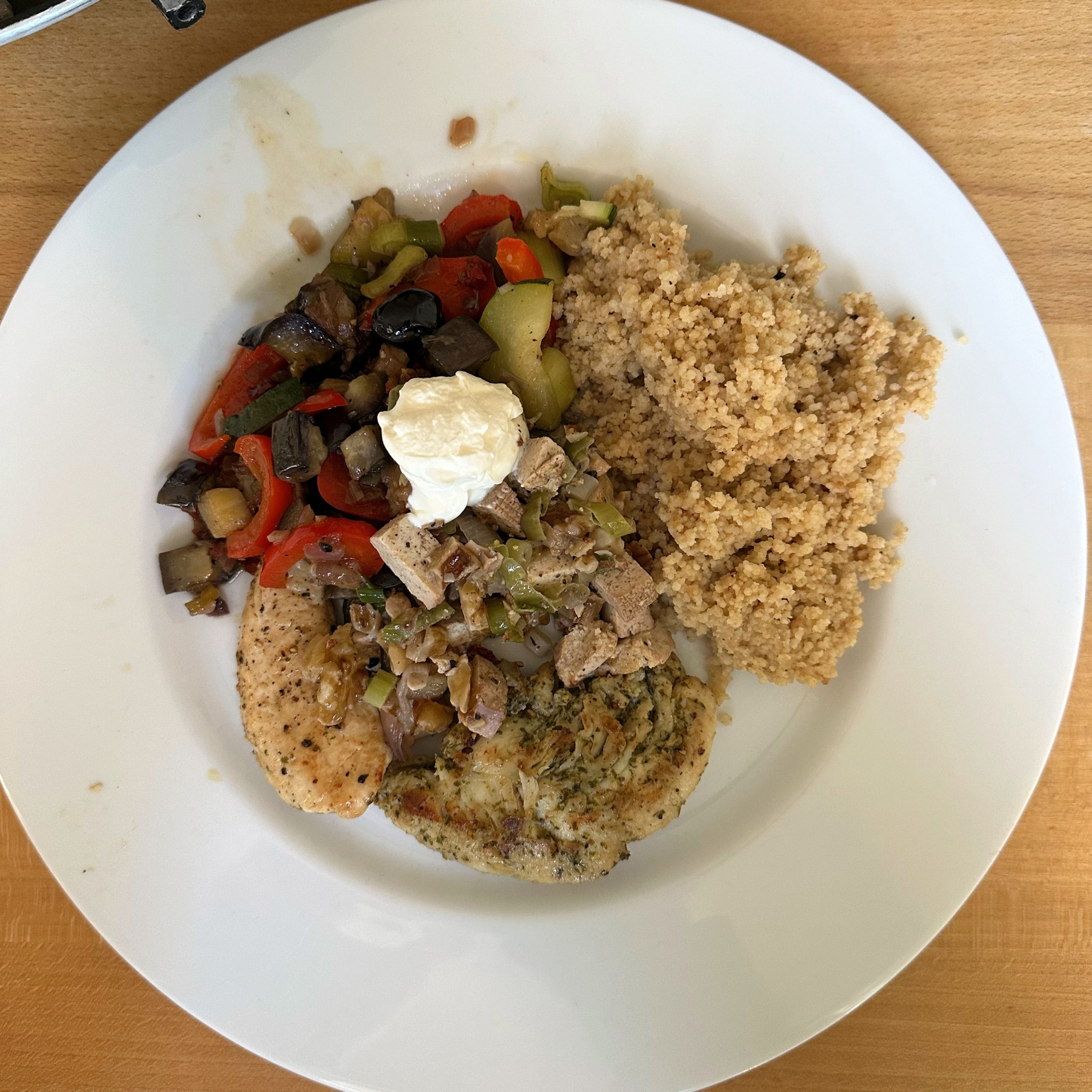 Teller mit zweierlei Hähnchenbrust (schlicht gewürzt und mit Kräuterkruste), belegt mit gebratenen Zwiebeln, Lauch und einem Klecks Joghurt, dazu Ratatouille aus Paprika, Aubergine, Zucchini und schwarzen Oliven sowie Couscous.