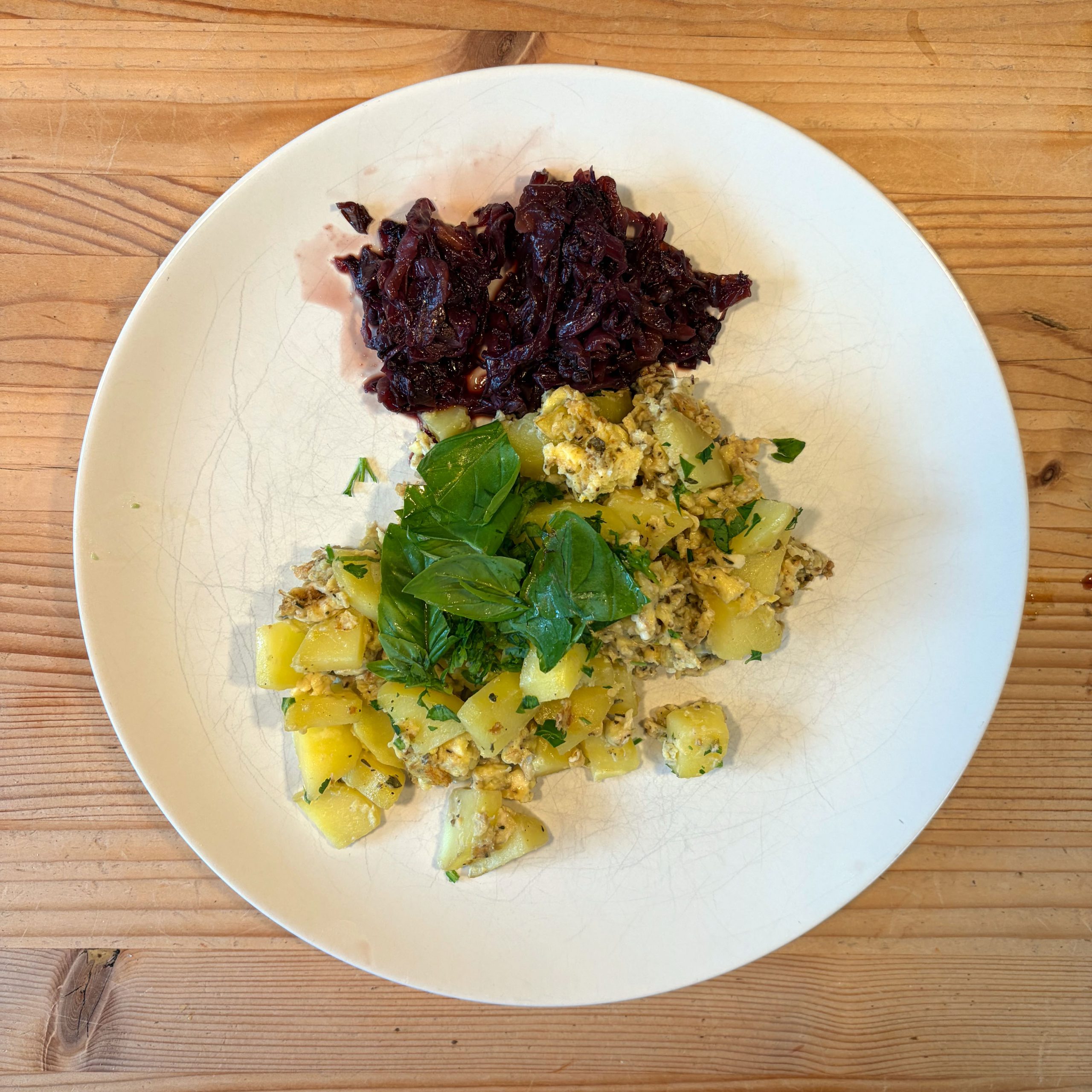 Weißer Teller mit zwei Komponenten: gebratene Kartoffelwürfel mit Spiegelei und frischen Kräutern (Basilikum, Petersilie) sowie geschmorter Radicchio, serviert auf einem Holztisch.
