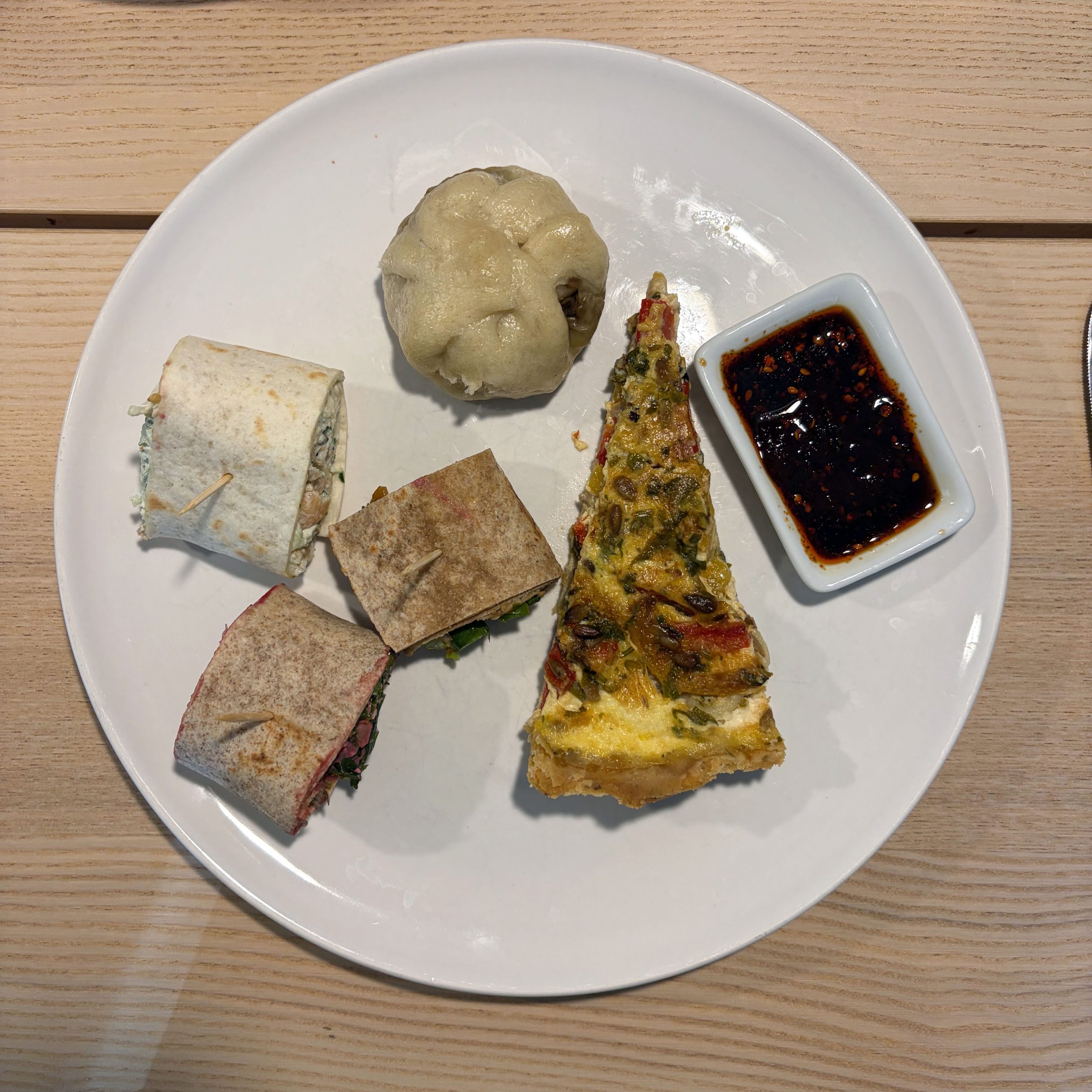 Ein weißer Teller mit einem Buffet-Teller-Arrangement: ein Stück Paprika-Tarte mit Ziegenfrischkäse und Sonnenblumenkernen auf Mürbeteig, ein gedämpftes Hefeklößchen (Bao), zwei halbierte Wraps mit Füllung, zwei halbierte Vollkornbrötchen-Sandwiches sowie ein kleines Schälchen mit dunkler Chilisoße.