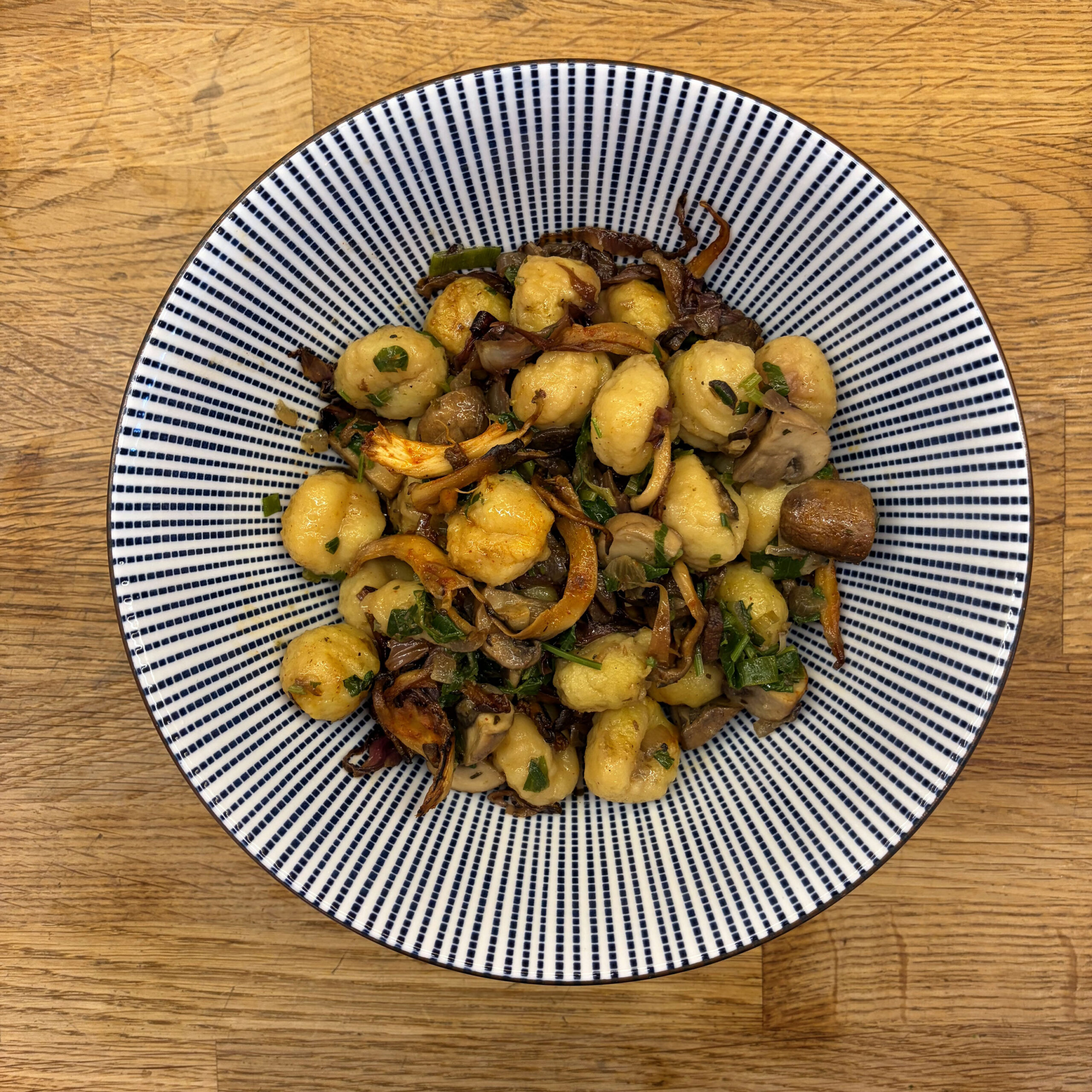 Gebratene Gnocchi mit karamellisierten Zwiebeln, Champignons, Austernpilzen und frischen Kräutern in einer blau-weiß gestreiften Schüssel auf einem Holztisch, Draufsicht.