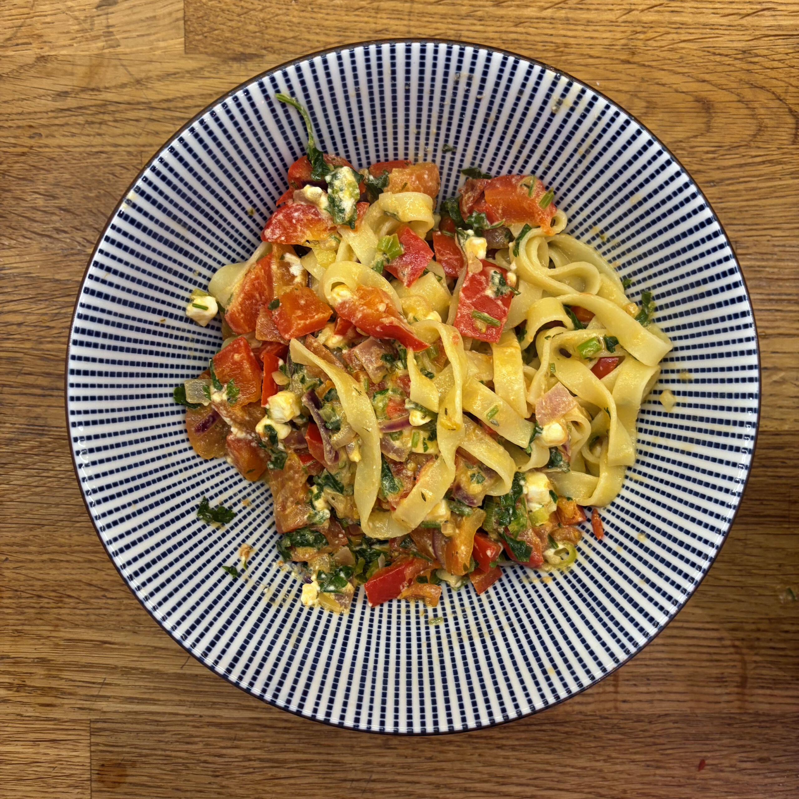 Tagliatelle mit Paprika-Tomatenmark-Soße, garniert mit zerbröseltem Feta und gehackter Petersilie, serviert in einer blau-weiß gestreiften Schüssel auf einem Holzuntergrund.