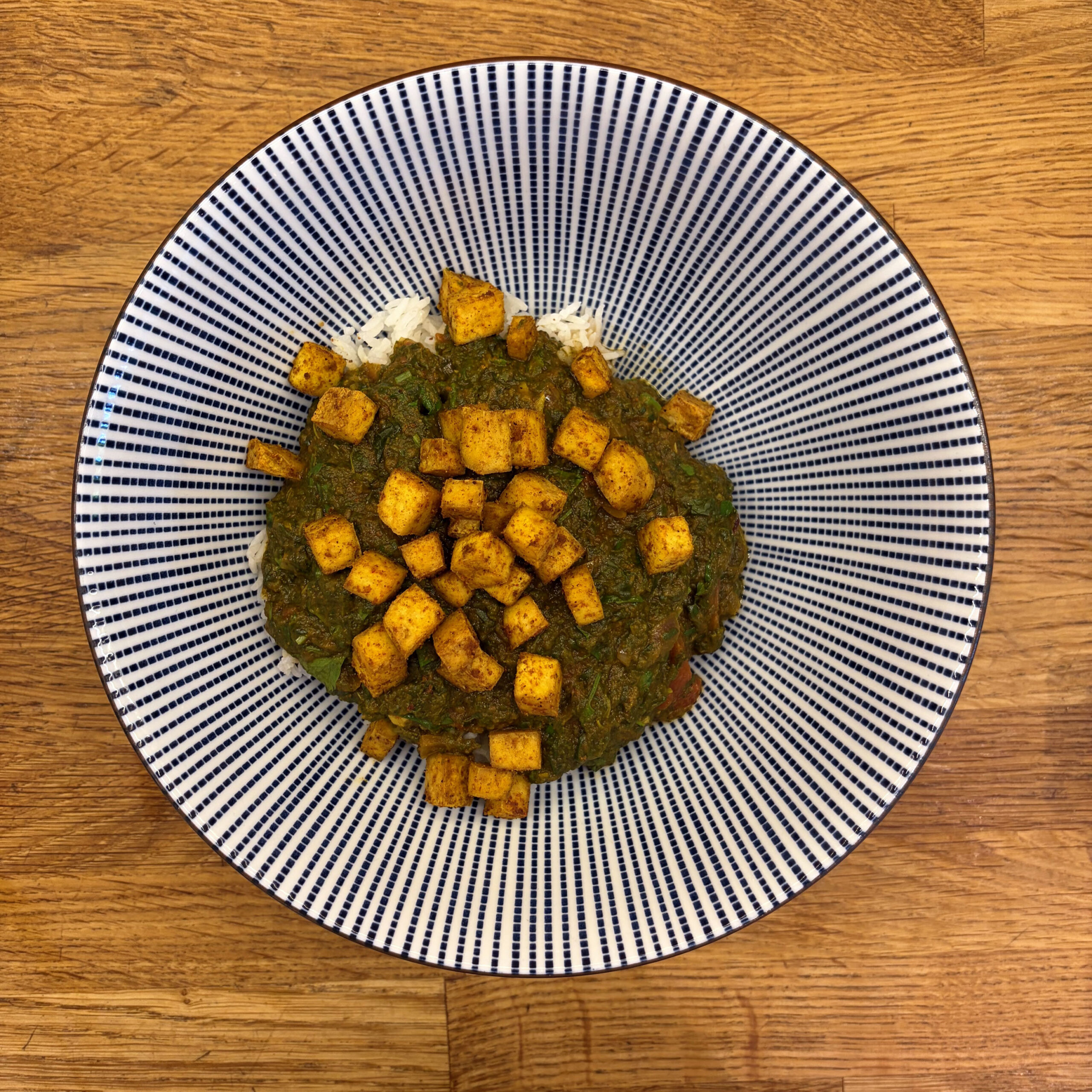 Teller mit Palak Tofu: gewürfelter, goldgelb gerösteter Tofu auf dunkelgrüner Spinat-Tomaten-Sauce, serviert mit weißem Basmatireis, auf einem blau-weiß gestreiften Teller.