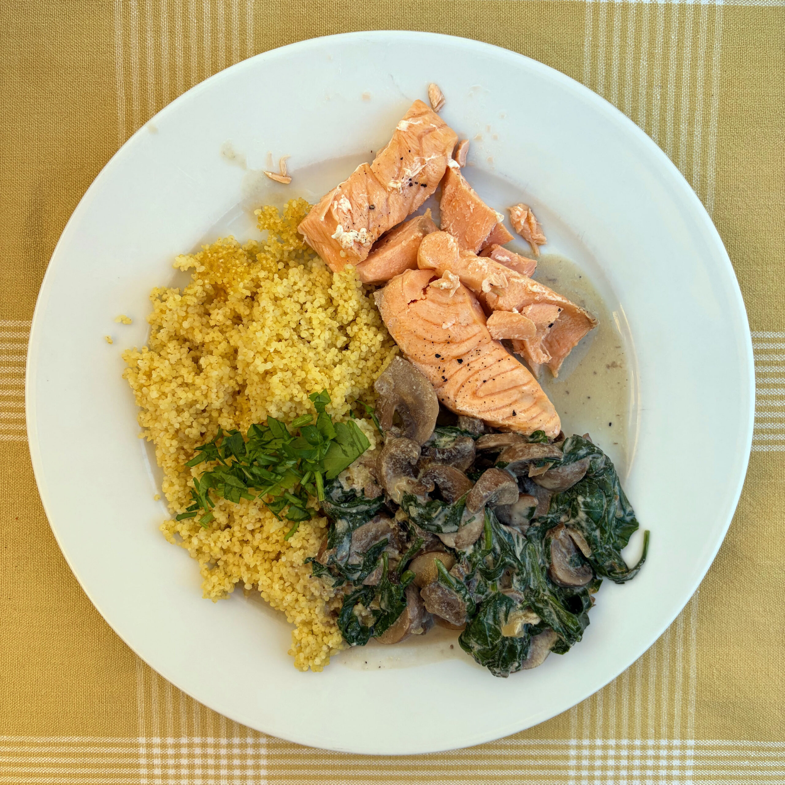 Teller mit gedämpftem Lachsfilet, gelblicher Hirse mit frischen Kräutern und cremiger Pilz-Spinat-Soße mit Champignons auf gelbem Karostoff.