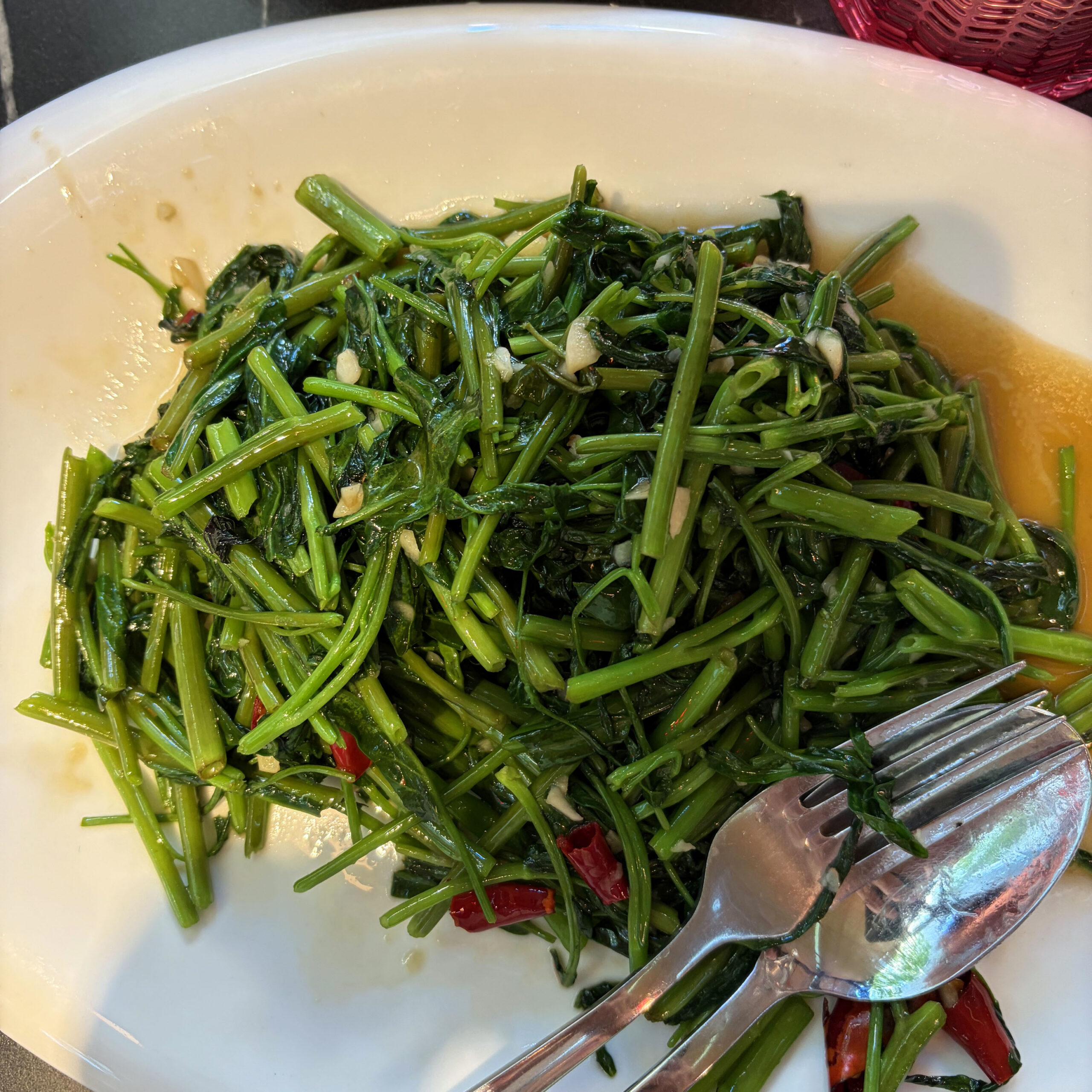 Stir-fried Water Spinach (Kangkung) mit Knoblauch und roten Chilischoten auf einem weißen Teller, serviert mit Gabel und Löffel – ein typisches Gericht der Szechuan-Küche.