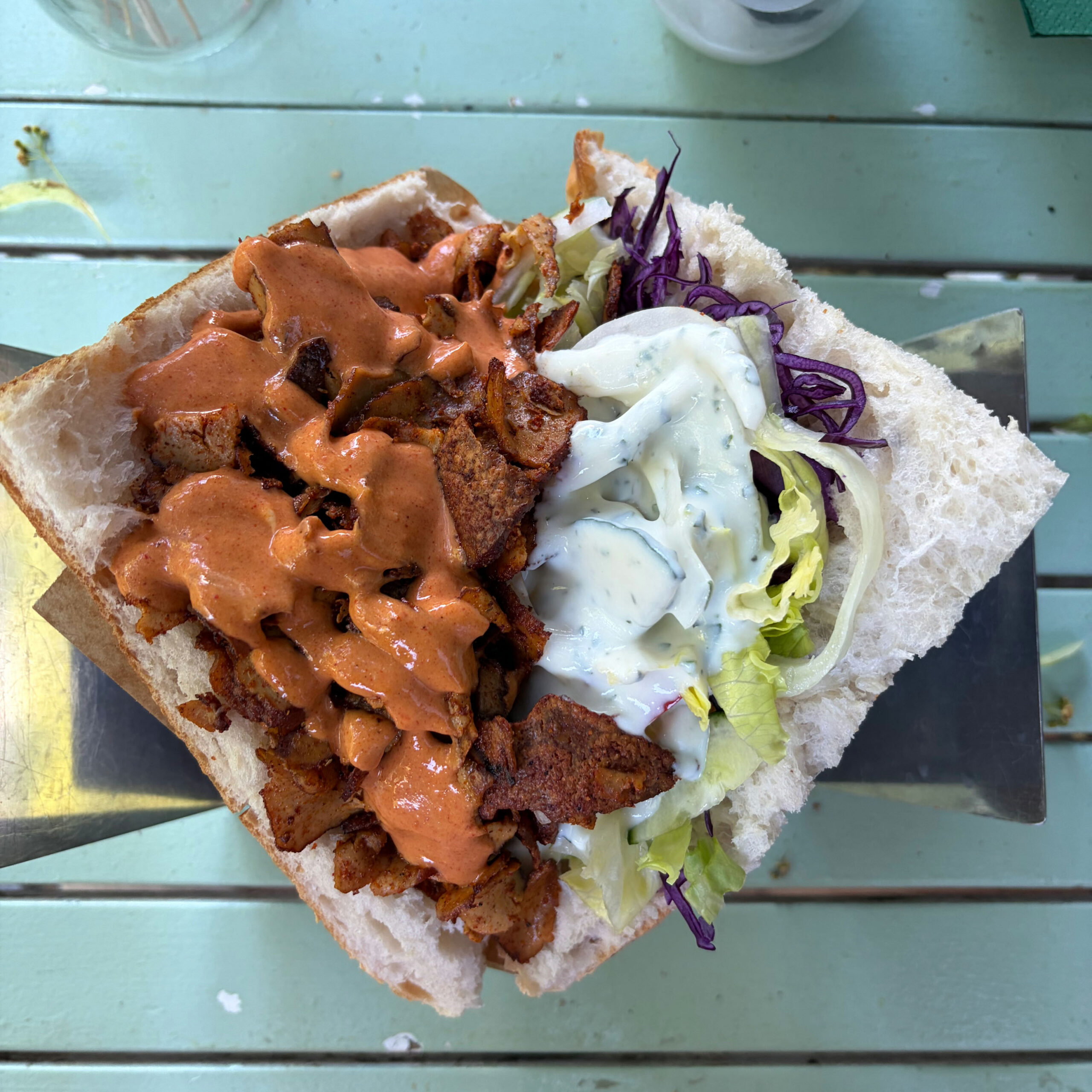Veganer Döner (Fiöner) von Vamos Veganos in einem Weißbrotbrötchen, belegt mit gewürztem Ersatzfleisch und roter Soße auf der linken Seite sowie Eisbergsalat, Rotkohl und Knoblauchsoße auf der rechten Seite, von oben fotografiert auf einem mintgrünen Metalltisch.