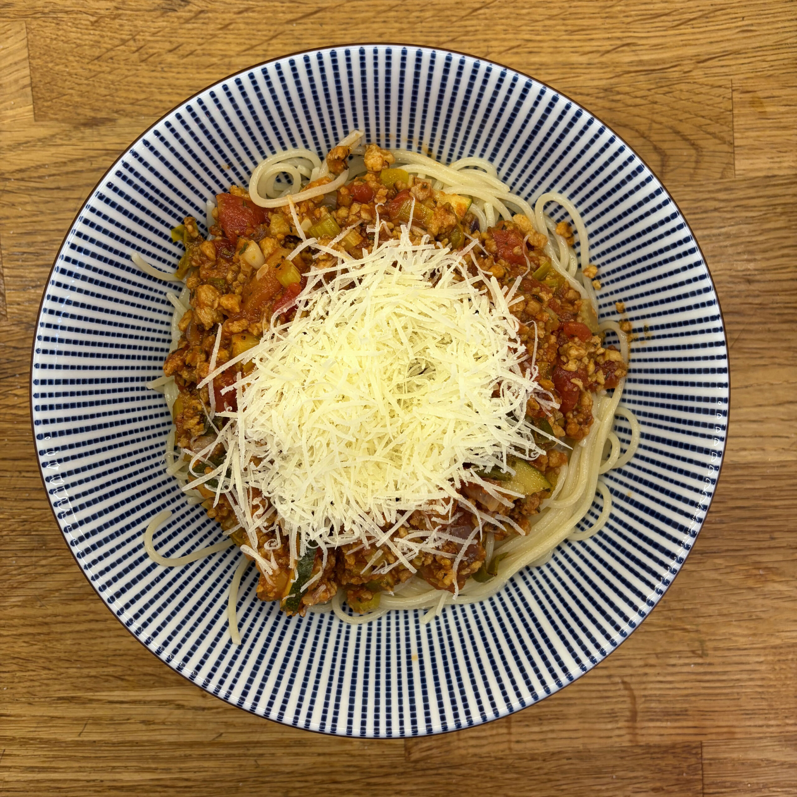 Eine Portion Spaghetti Bolognese mit Sojahack in einer blau-weiß gestreiften Schüssel, angerichtet auf einem Holztisch. Die Nudeln sind mit einer Tomatensauce aus Sojahack und Gemüse bedeckt und mit einem großzügigen Berg frisch geriebenem Parmesan garniert.
