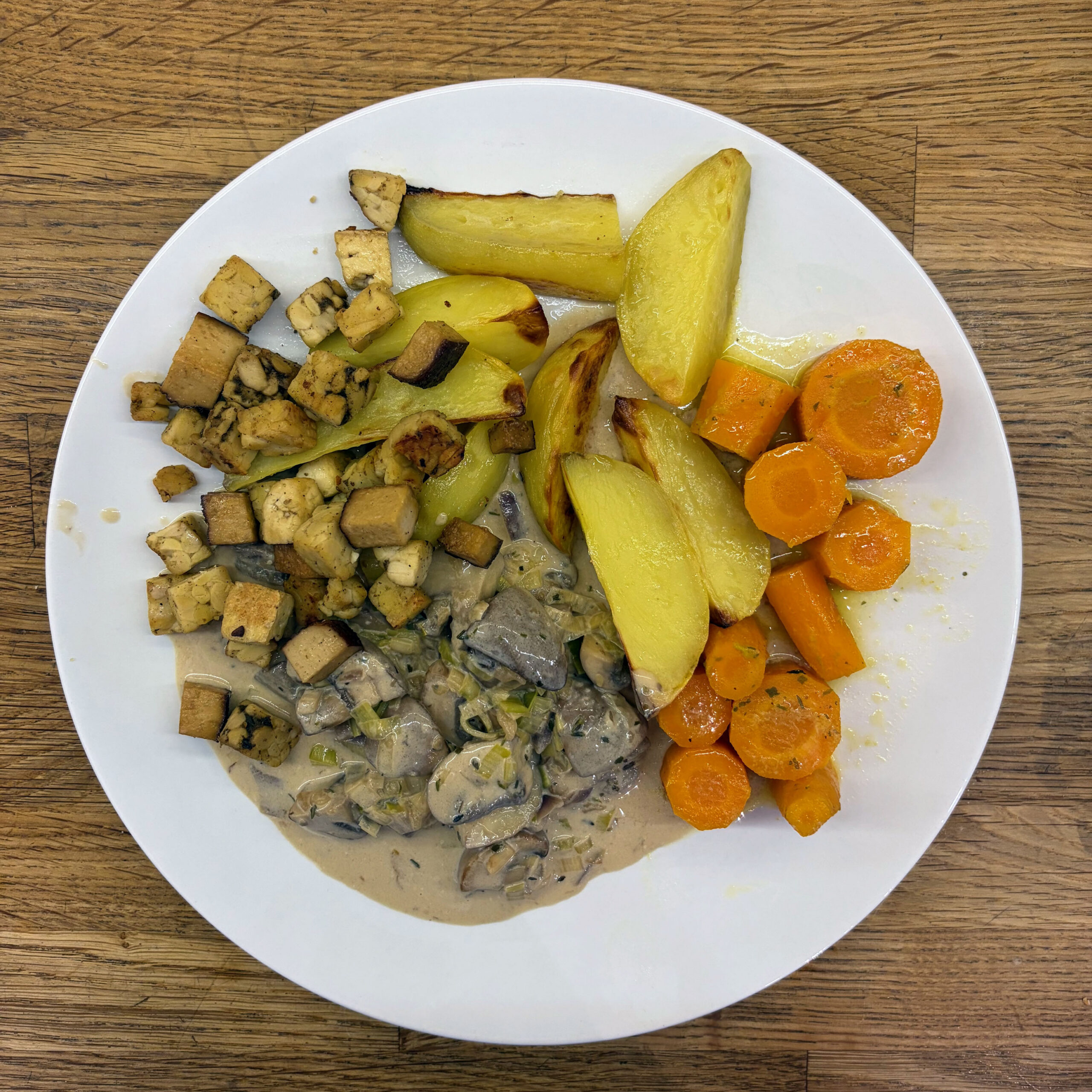Teller mit Champignon-Lauch-Rahmsauce in Mandelsahne, gebratenem Tempeh in Würfeln, glasierten Möhrenscheiben und goldbraunen Ofenkartoffelspalten.