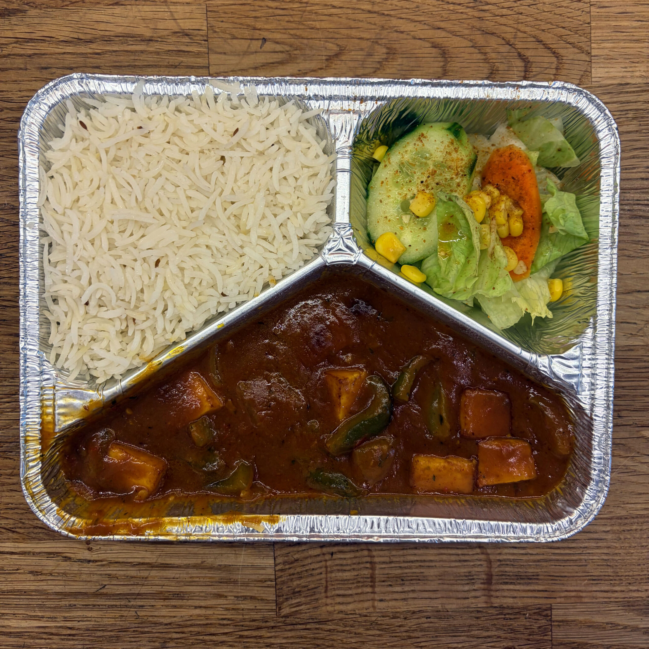 Dreigeteilte Aluminiumschale mit Chili Paneer (Käsewürfel in roter Currysauce mit grünen Paprikastücken), weißem Basmati-Reis und einem Beilagensalat mit Gurke, Karotte, Mais und Eisbergsalat, auf Holzuntergrund.