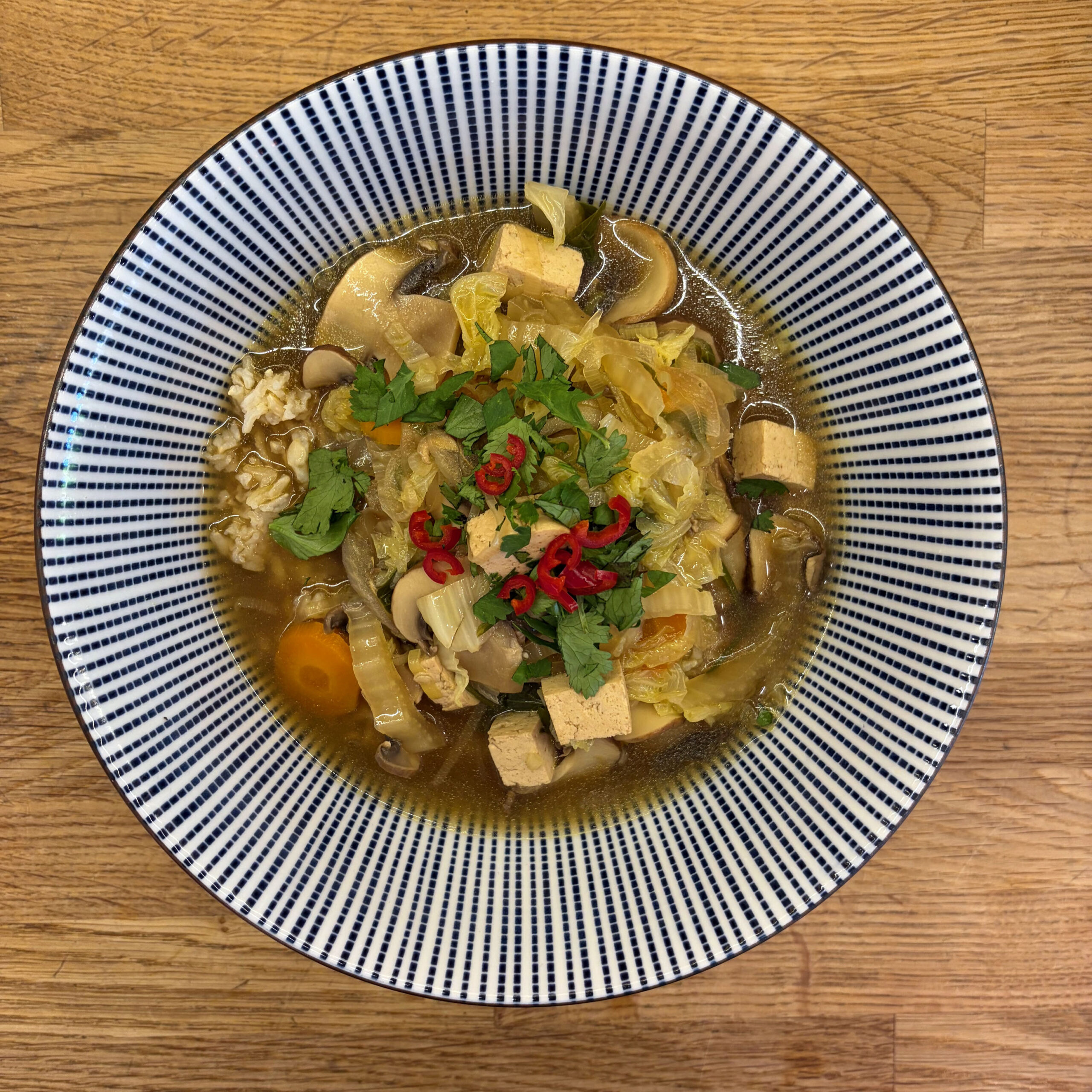 Vegane Pho in einer blau-weiß gestreiften Schüssel: klare Brühe mit Tofuwürfeln, Champignons, Chinakohl und Ei, garniert mit frischem Koriander und roten Chilischoten.