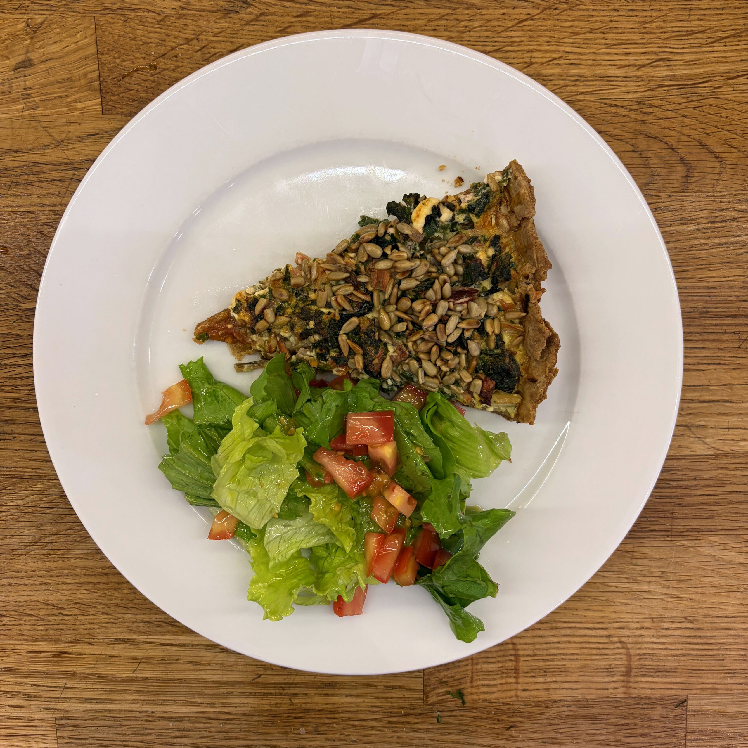 Ein Stück Mangoldtarte mit Spinat, Feta und gerösteten Sonnenblumenkernen auf einem Mürbeteigboden, serviert mit einem grünen Blattsalat mit Tomatenstücken und Vinaigrette, angerichtet auf einem weißen Teller.