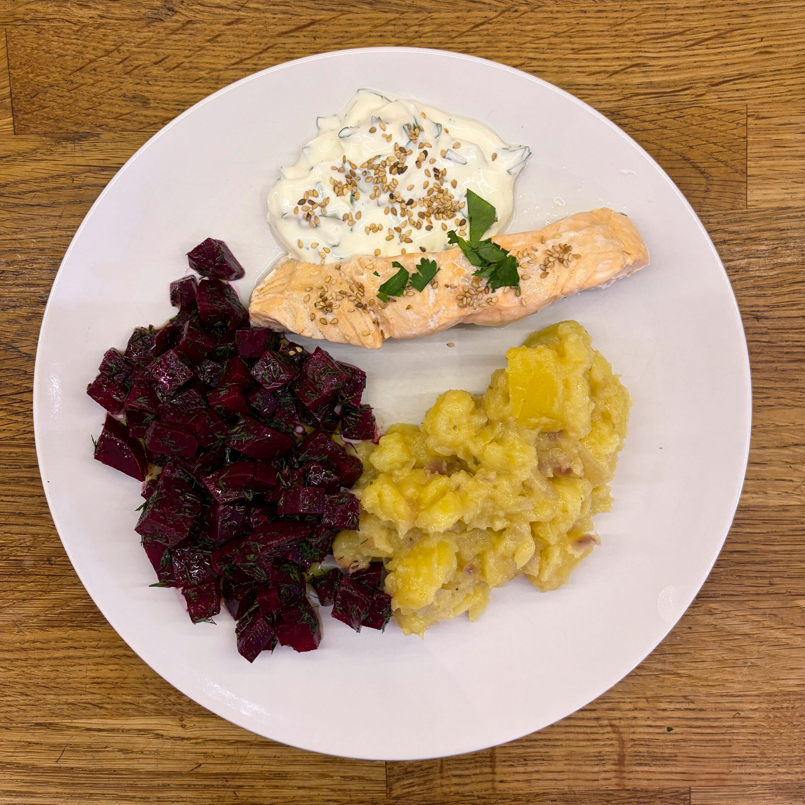 Teller mit gedämpftem Lachsfilet, garniert mit Koriander und Sesam, serviert mit Joghurt-Koriander-Dipp, gewürfeltem Rote-Bete-Salat mit Dill sowie warmem Kartoffelsalat.