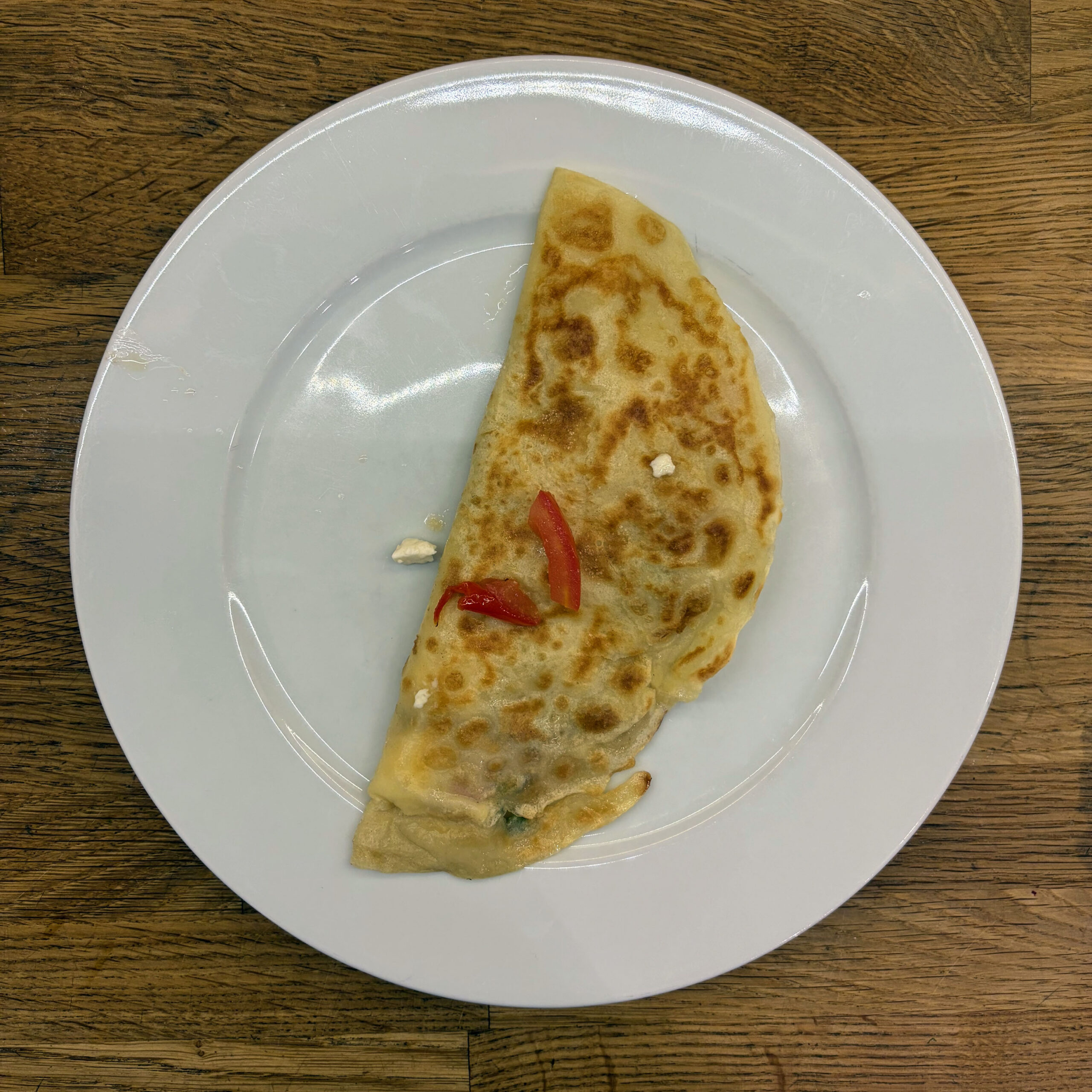 Goldbraun gebratener, halbmondförmig gefalteter Pfannkuchen mit Feta-Füllung und roten Paprikastreifen auf weißem Teller, von oben fotografiert.