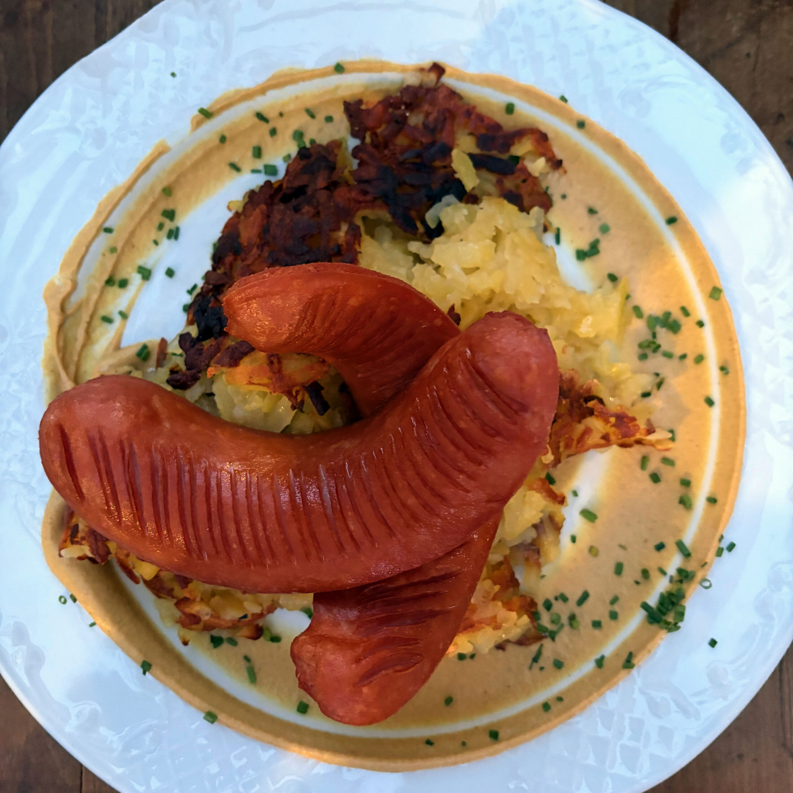 Zwei gegrillte Augsburger Würste auf einem Rösti mit Sauerkraut, angerichtet auf einem weißen Teller mit cremiger Senfsauce und frischen Schnittlauchröllchen.