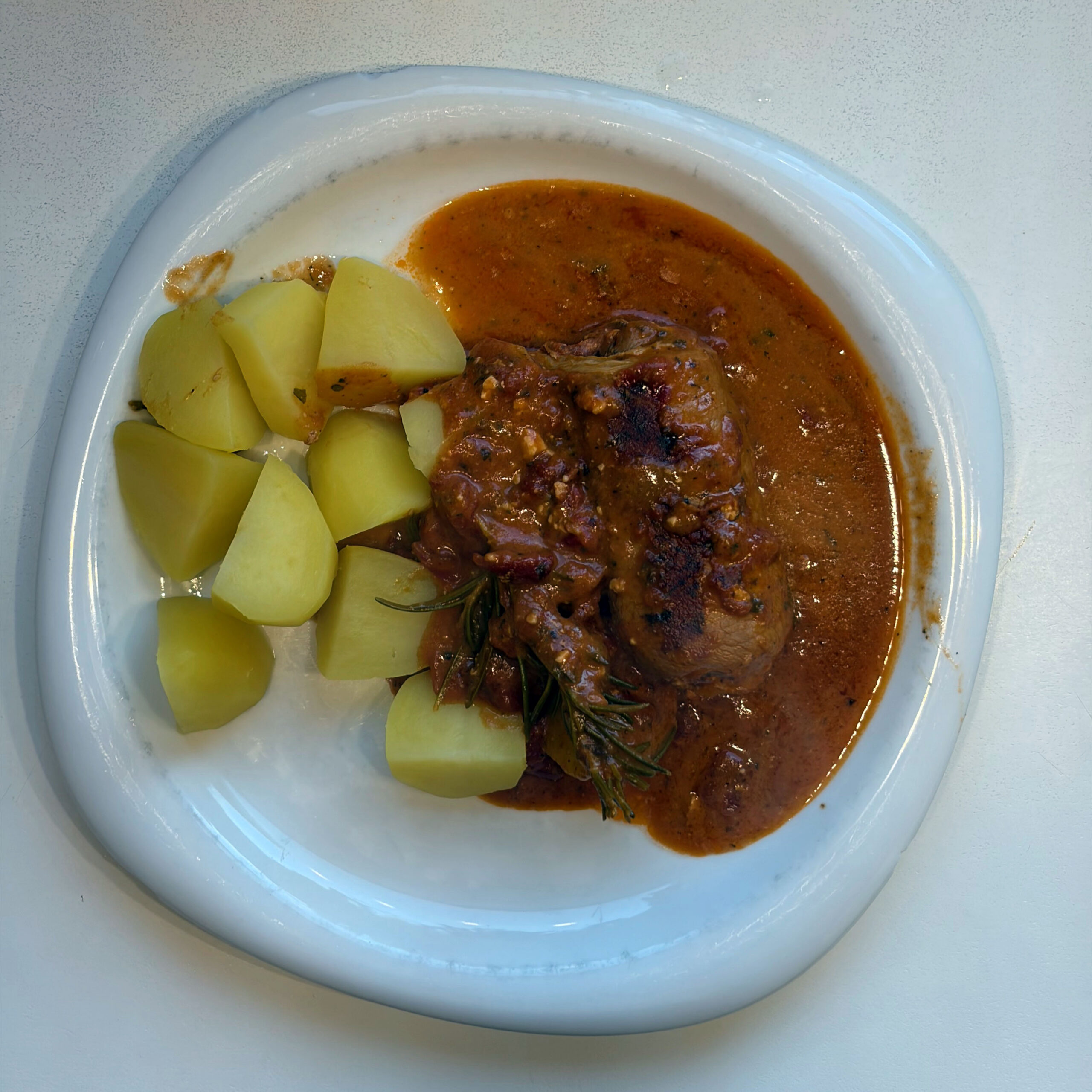 Ein weißer Teller mit einer Rindsroulade griechischer Art in einer kräftigen Tomatensauce mit Rosmarin, serviert mit gekochten Salzkartoffelwürfeln.