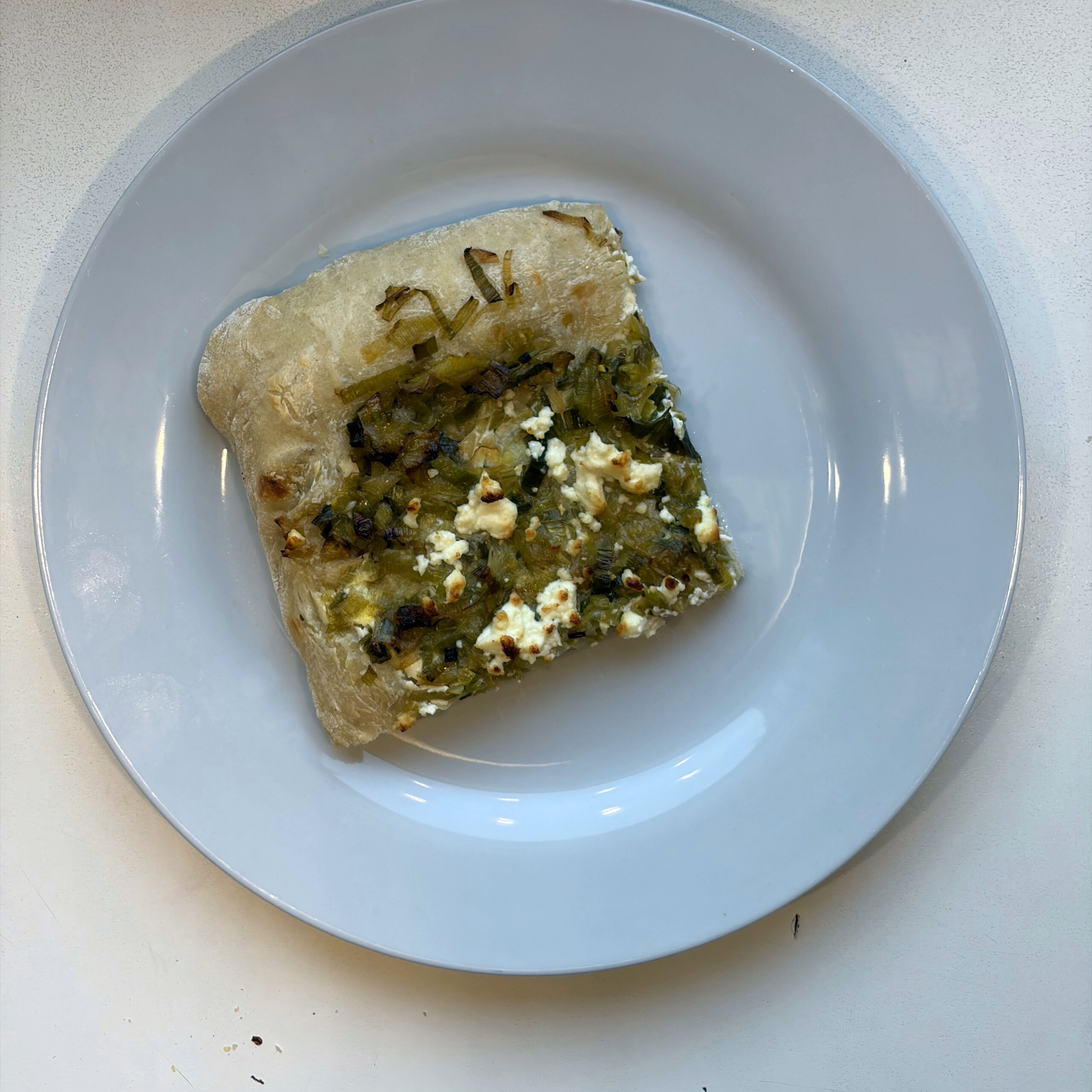 Ein Stück Flammkuchen mit karamellisiertem Lauch und zerbröseltem Feta auf einem hellgrauen Teller, serviert auf hellem Untergrund.