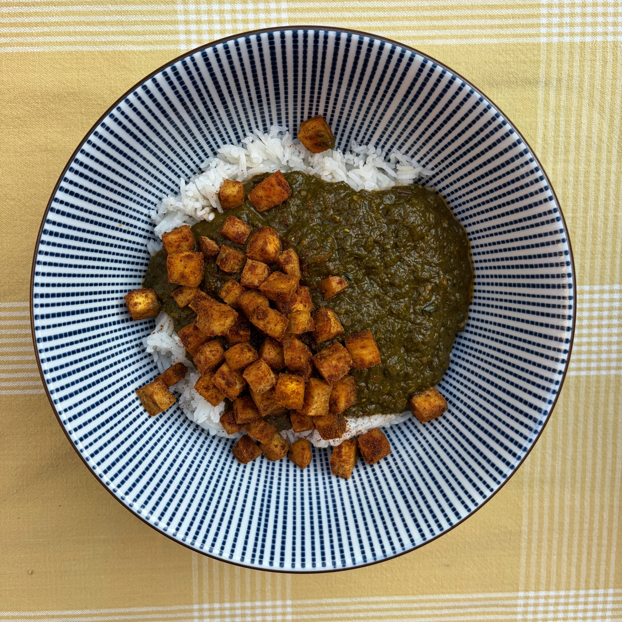 Ein Teller Palak Tofu: weißer Basmatireis, belegt mit dunkelgrüner Spinatsauce und goldbraun gerösteten, gewürzten Tofuwürfeln, serviert in einer blau-weiß gestreiften Schale auf gelbem Karostoff.