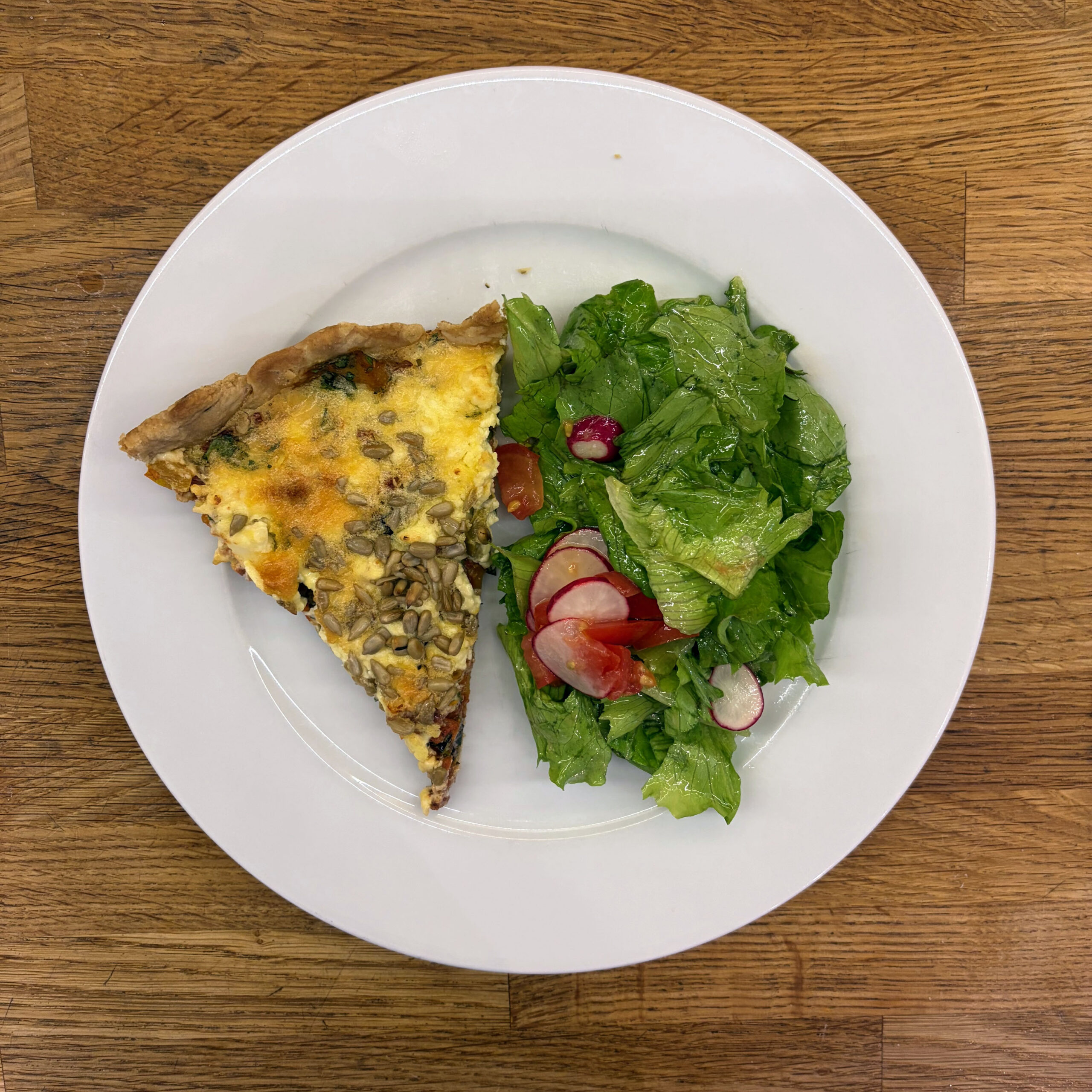 Ein Stück Paprika-Feta-Tarte mit Mürbeteigboden, belegt mit gerösteten Paprikastreifen, Oliven, getrockneten Tomaten, zerbröseltem Feta und gerösteten Sonnenblumenkernen, serviert auf einem weißen Teller neben einem grünen Blattsalat mit Radieschen und Tomaten.
