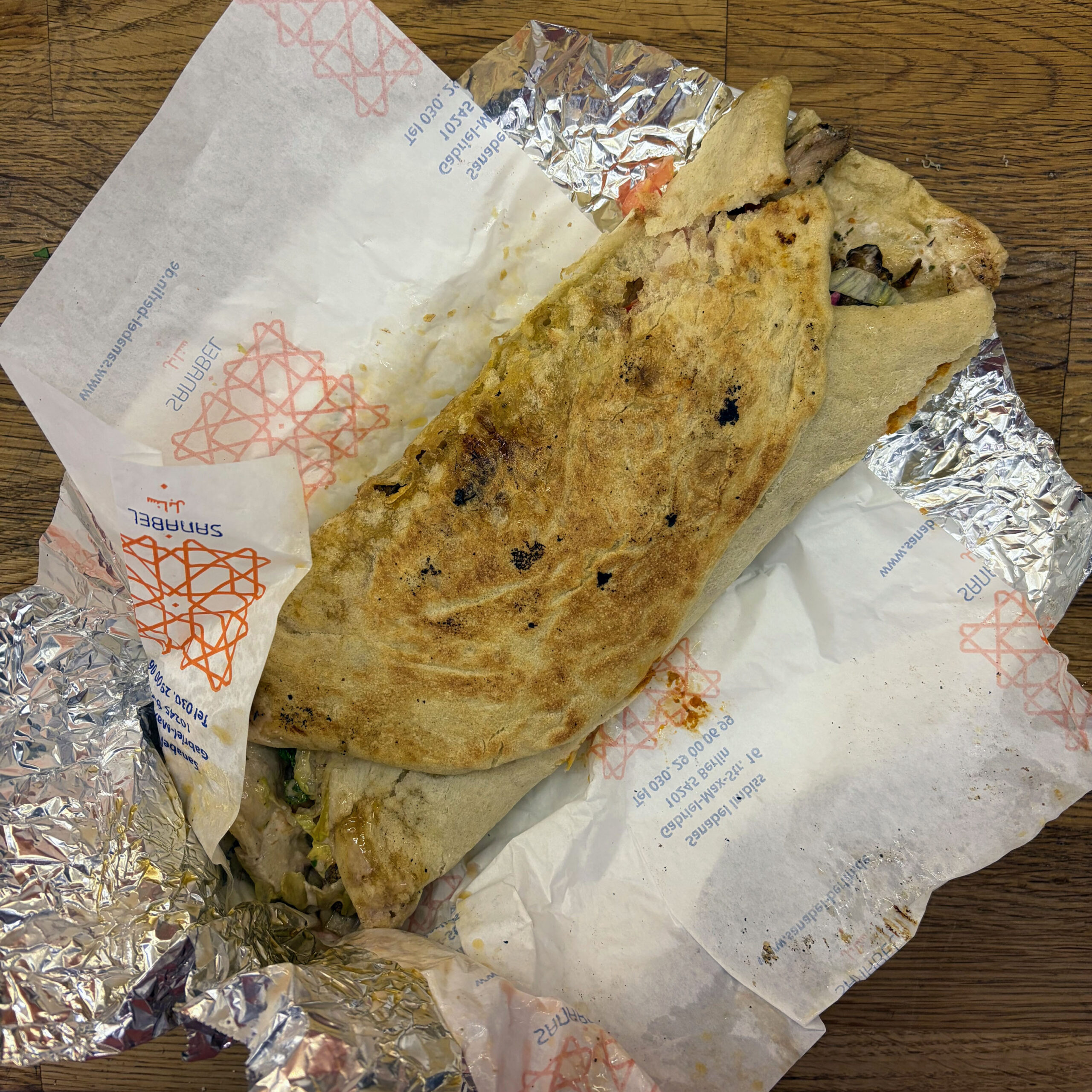 Aufgeklapptes Shawarma-Sandwich vom Berliner Restaurant Sanabel, serviert auf Einwickelpapier mit dem charakteristischen roten Sanabel-Logo und Alufolie, auf einem Holztisch.