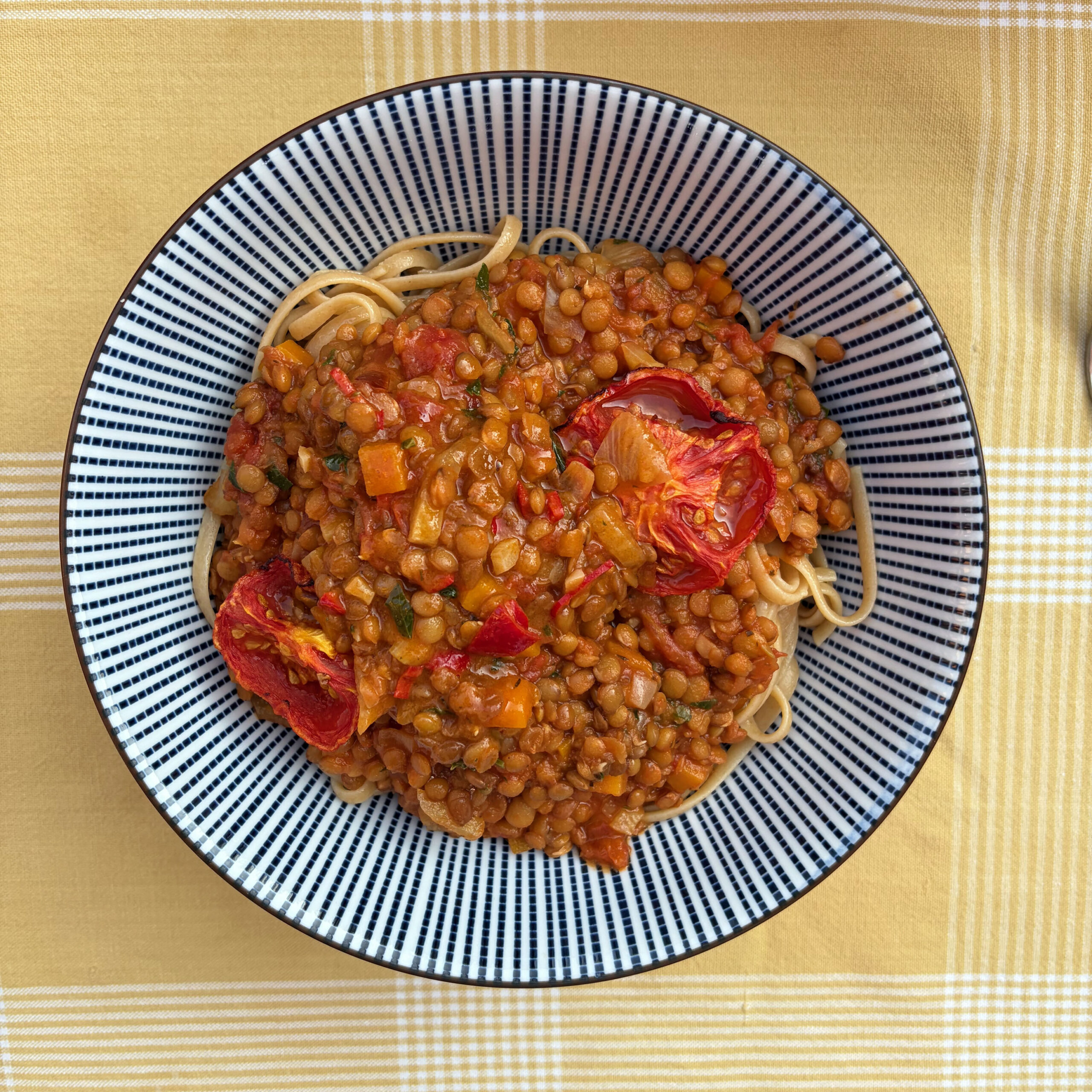 Schüssel mit Vollkorn-Spaghetti, belegt mit Linsenbolognese aus Tellerlinsen und Tomatensauce, garniert mit gerösteten Ofentomaten, auf gelbem Karostoff.