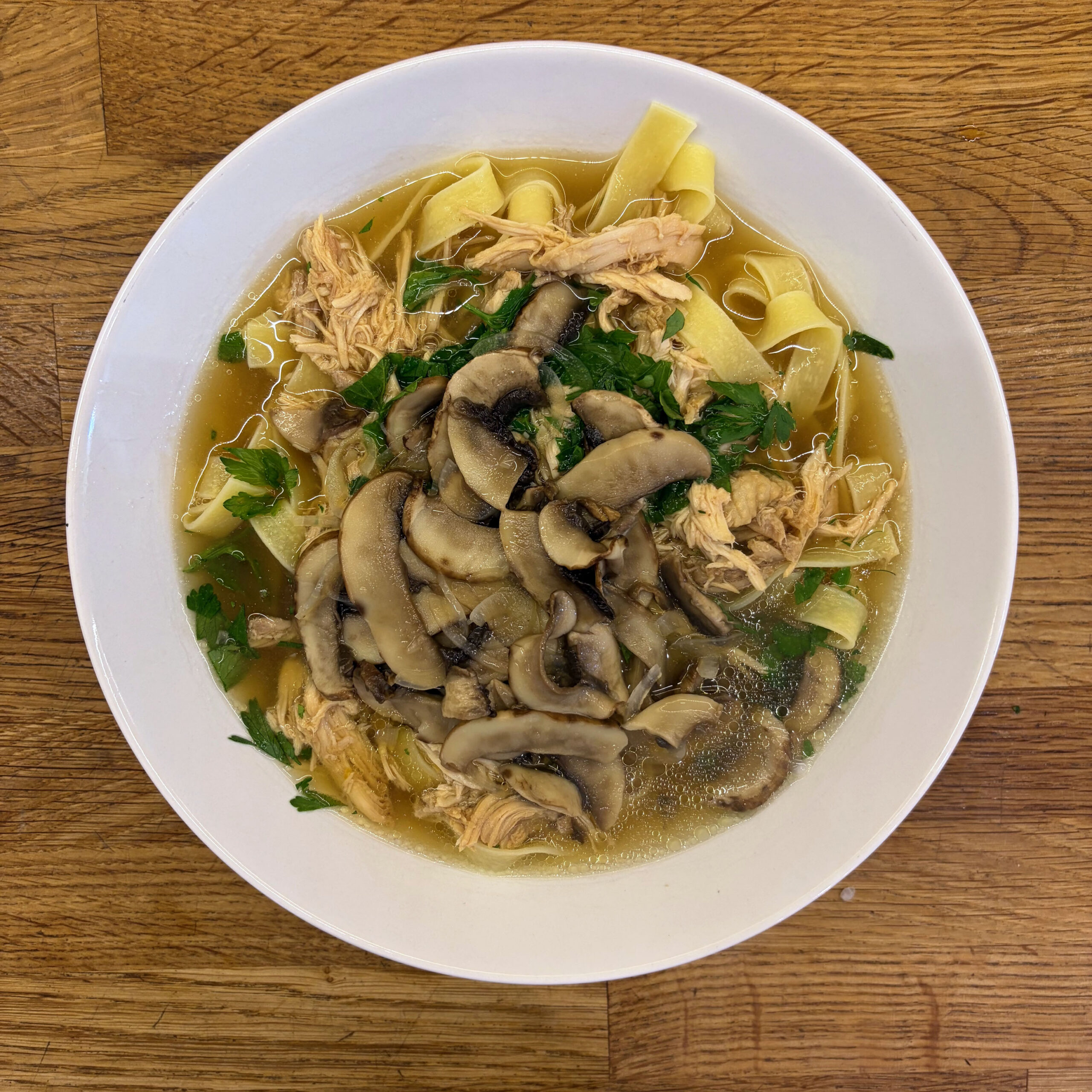 Hühnersuppe mit breiten Tagliatelle-Nudeln, zerzupftem Hühnerfleisch, reichlich Champignonscheiben und frischer Petersilie in goldgelber Brühe, serviert in einem weißen tiefen Teller auf einer Holzoberfläche.
