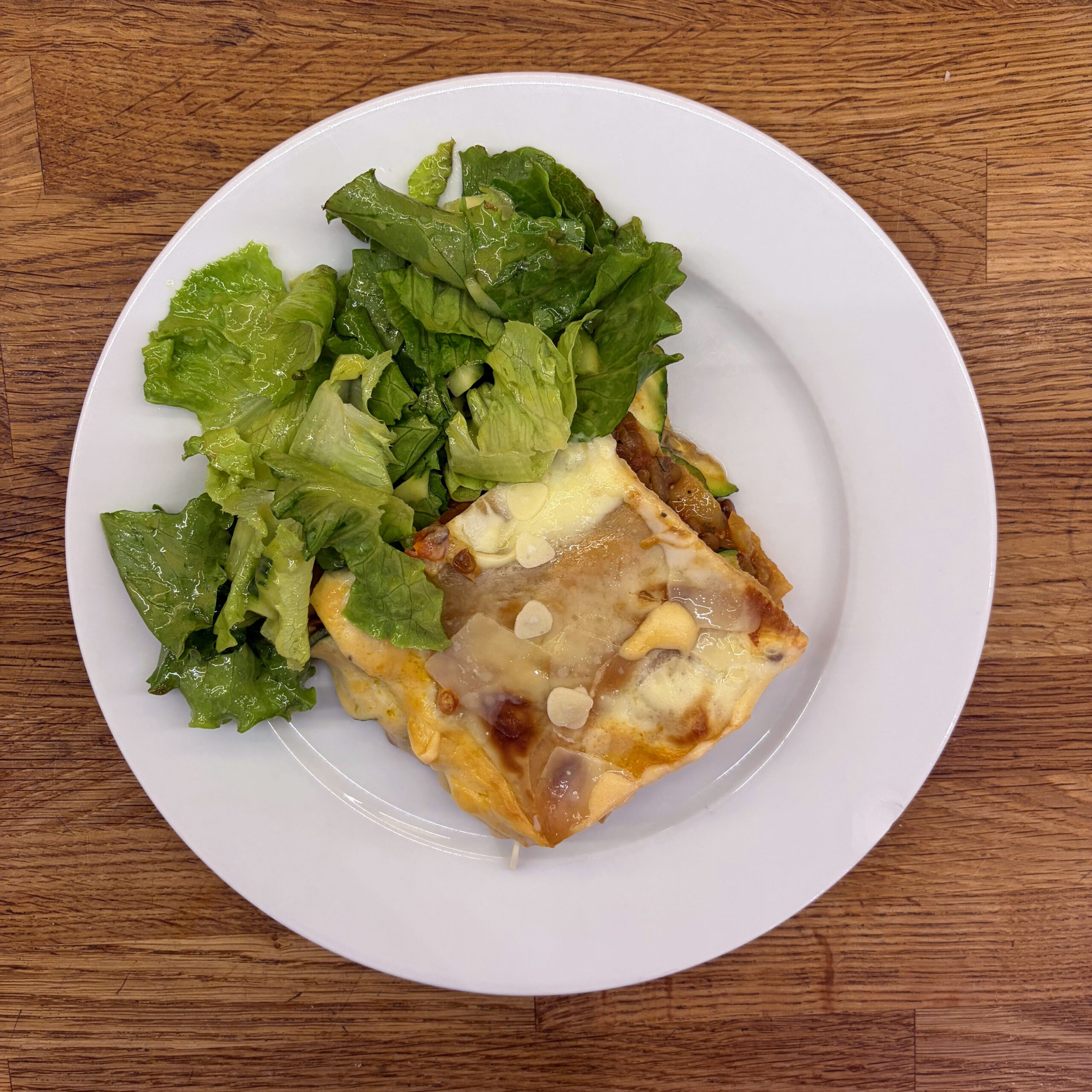 Ein Stück Linsen-Zucchini-Lasagne mit goldbrauner Béchamelkruste und gehobeltem Parmesan, serviert mit frischem grünem Blattsalat auf einem weißen Teller.