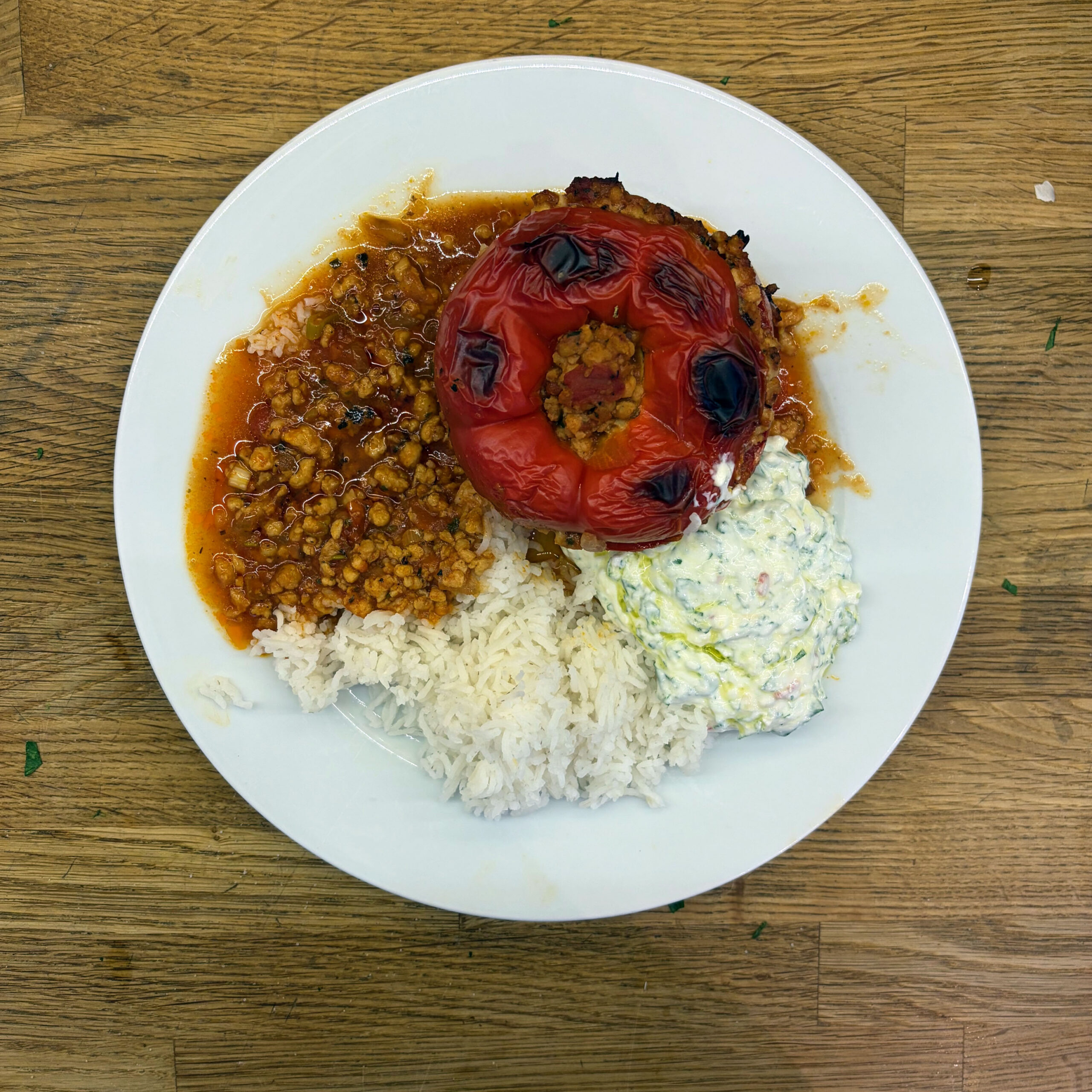Draufsicht auf einen weißen Teller mit einer gefüllten, gebackenen roten Paprika auf würziger Tomatensoße mit Sojagranulat, weißem Reis und einem cremigen Quark-Dip mit Kräutern.