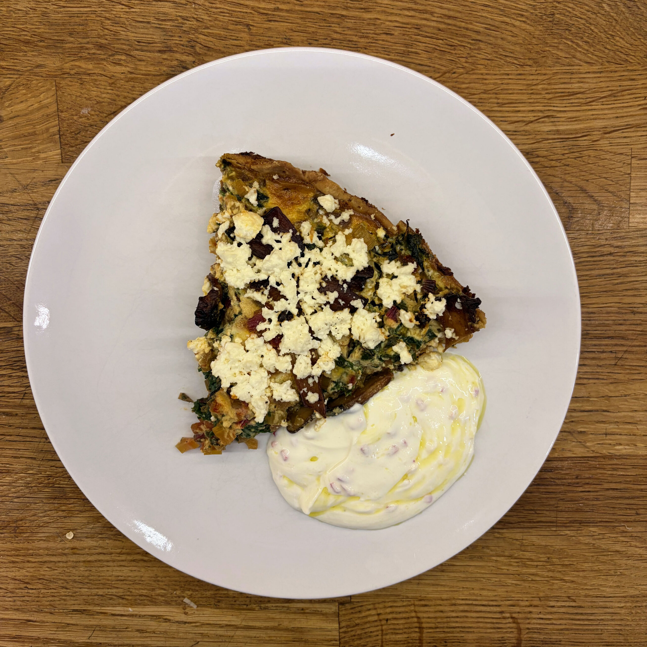 Ein Stück herzhafte Mangoldtarte mit gebratenen Kräuterseitlingen, belegt mit zerbröseltem Feta, serviert auf einem weißen Teller mit einem cremigen Joghurt-Quark-Dip, beträufelt mit Olivenöl, auf einer Holzunterlage.