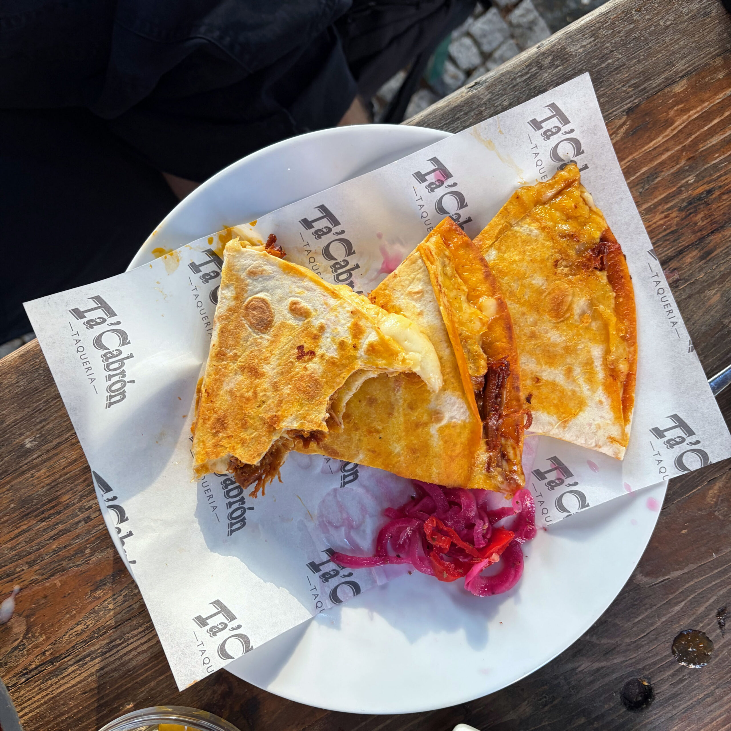 Zwei aufgeschnittene, goldbraun gegrillte Quesadillas mit geschmolzenem Käse und Fleischfüllung auf einem weißen Teller, serviert mit eingelegten roten Zwiebeln auf Einwickelpapier des Restaurants Ta'Cabrón Taqueria.
