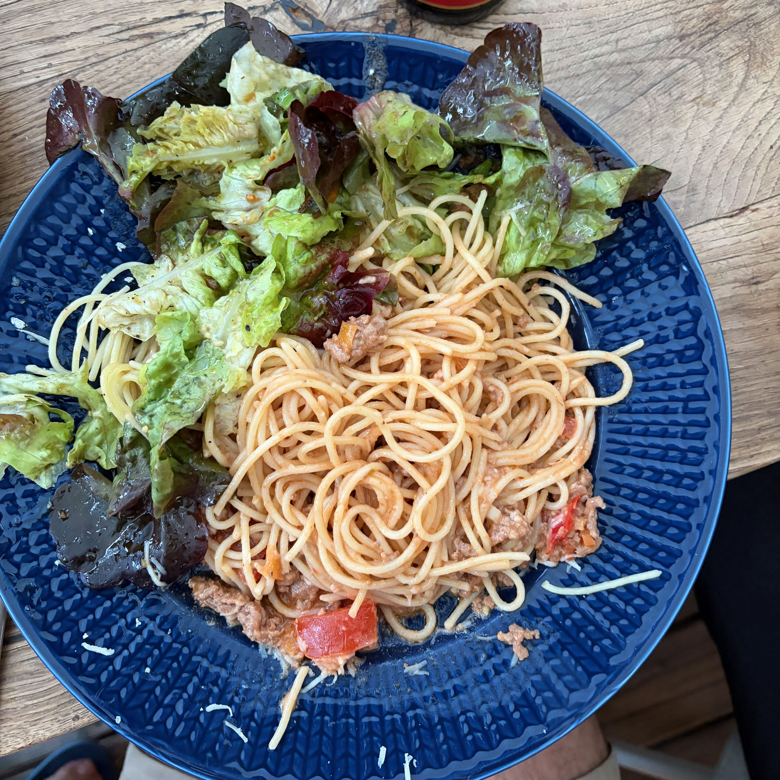Blauer tiefer Teller mit Spaghetti in Tomaten-Bolognese-Sugo mit Gemüsestücken und einer Beilage aus gemischtem grünen Salat mit roten Blättern, serviert auf einem Holztisch.