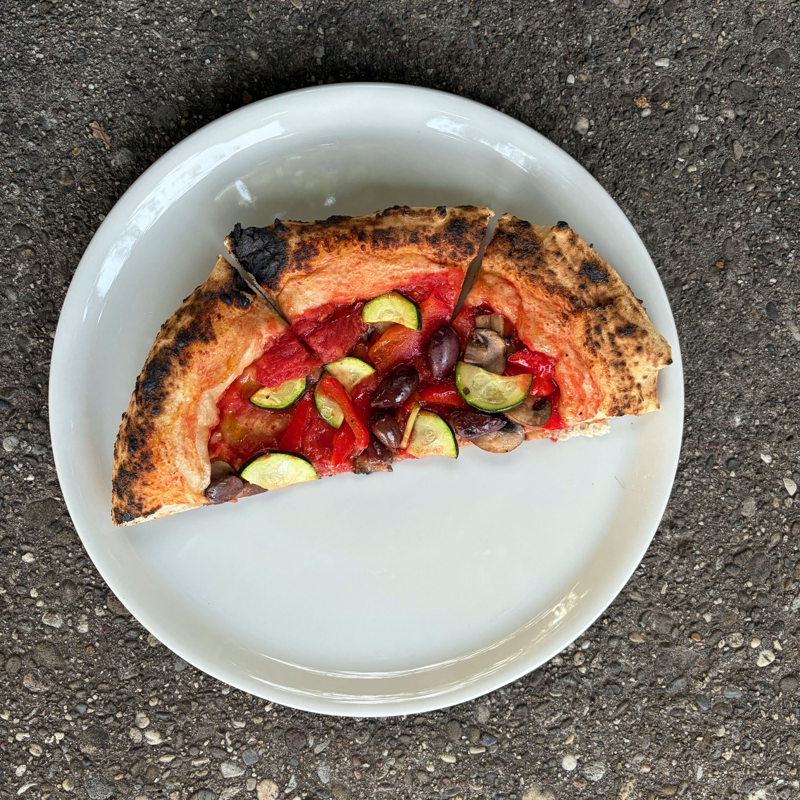 Ein Stück Neapolitanische Pizza mit Tomatensoße, Zucchini, roten Paprika, Kalamata-Oliven und Champignons auf einem weißen Teller. Der Rand ist im Holzofen charakteristisch dunkel gefleckt.