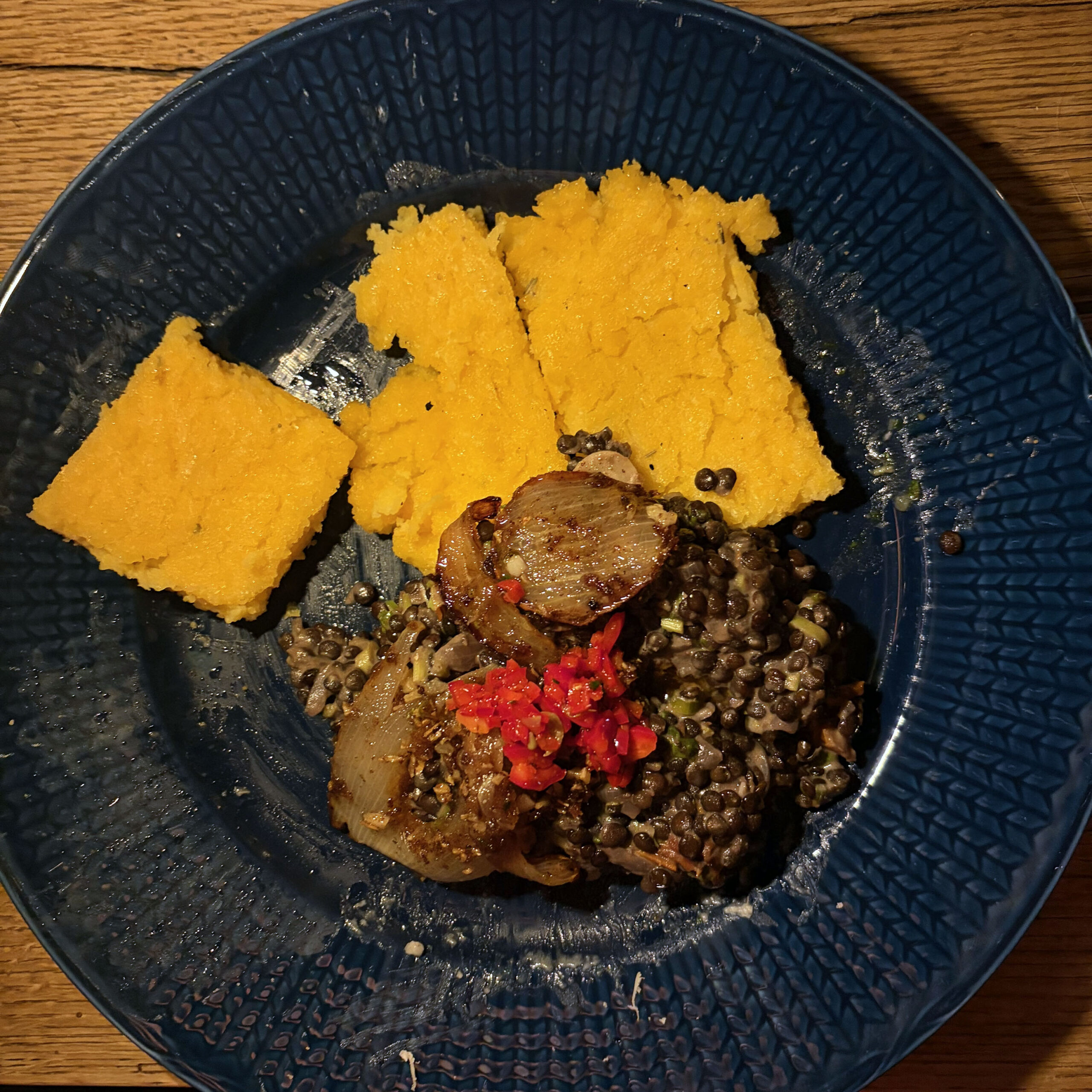 Blauer Teller mit drei Stücken gebackener goldgelber Polenta, Beluga-Linsen mit gebratenen Zwiebelscheiben und gehackten roten Chilischoten als Garnitur auf einem Holztisch.