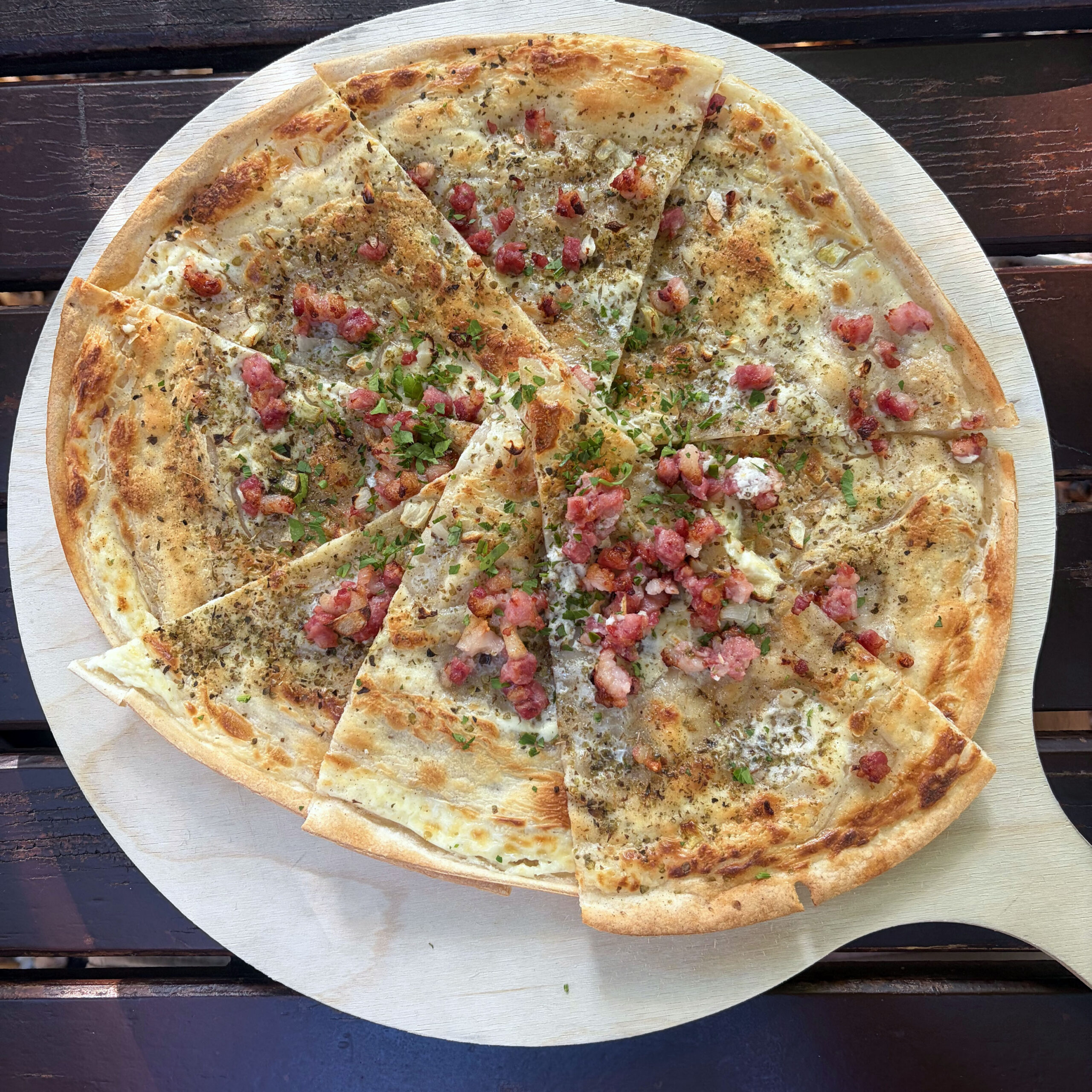 Klassischer Flammkuchen mit Speckwürfeln, Zwiebeln, Crème fraîche und frischen Kräutern auf einem runden Holzbrett, in mehrere Stücke geschnitten, serviert im Biergarten.