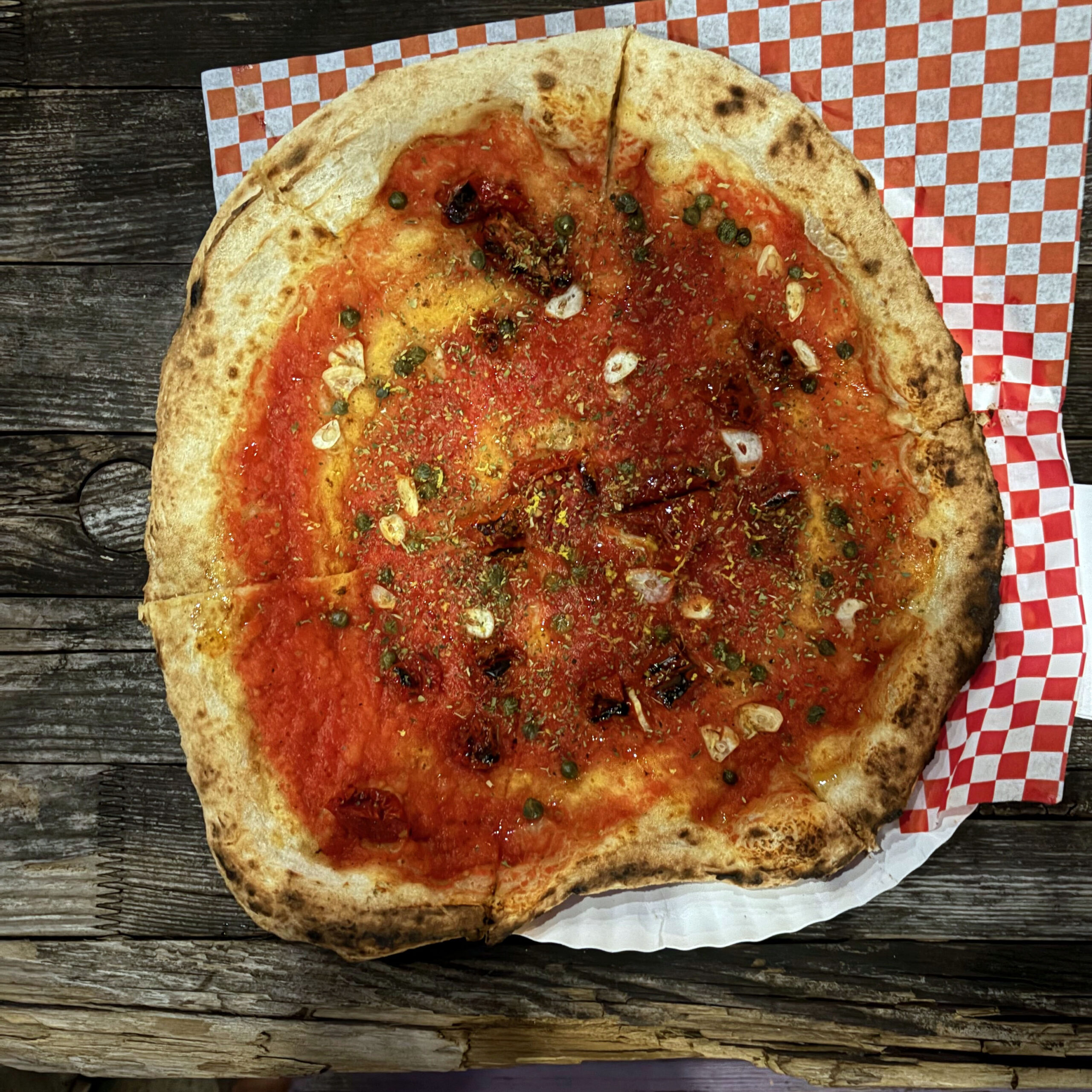 Pizza Marinara „Rime of the Ancient Marinara" von Slices of Doom: Neapolitanische Pizza mit charakteristisch geflecktem, luftigem Rand auf roter San-Marzano-Tomatensauce, belegt mit Knoblauchscheiben, Kapern, halbgetrockneten Tomaten, Zitronenabrieb und Oregano, serviert auf rot-weiß kariertem Papier auf einem rustikalen Holztisch.