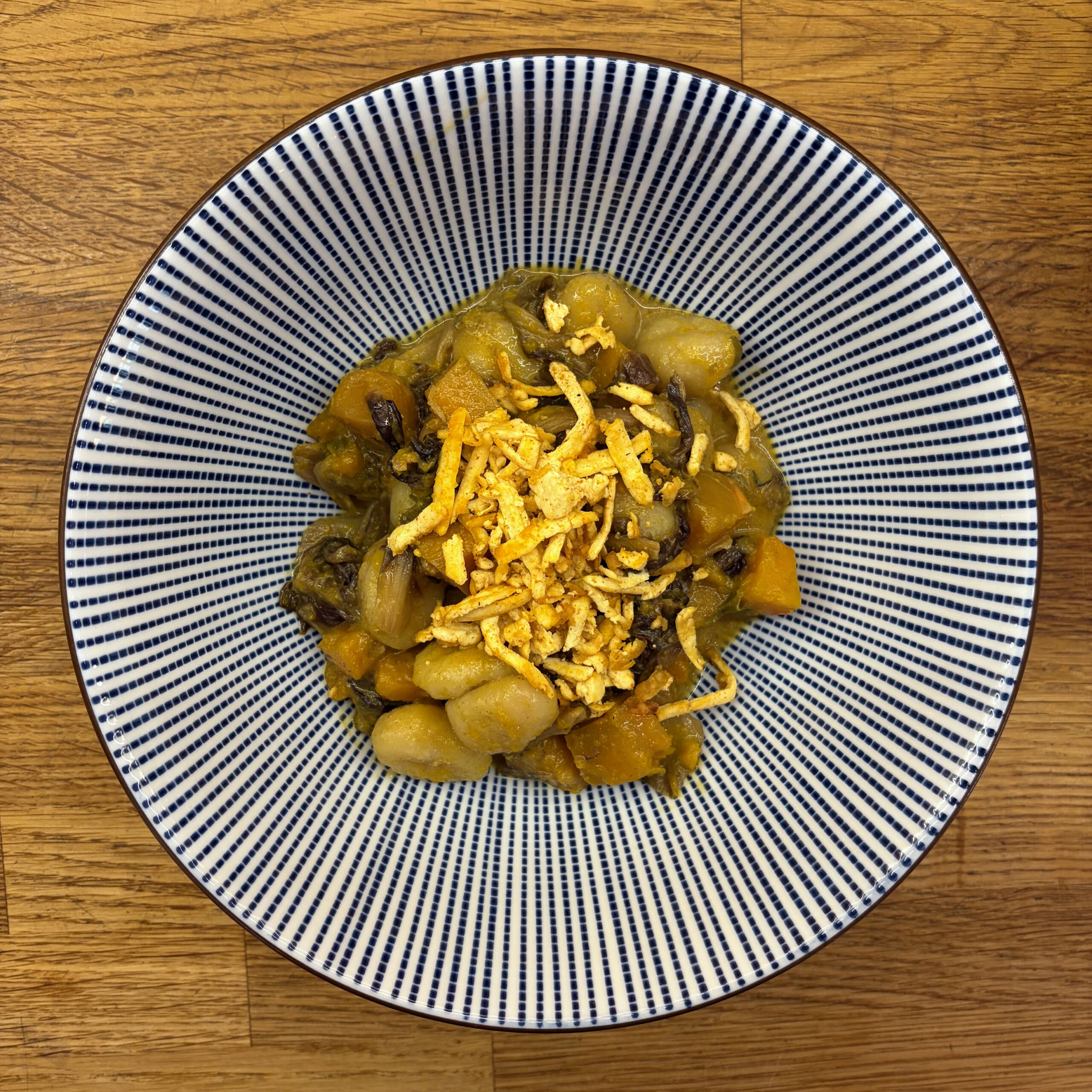 Gnocchi mit Kürbis-Radicchio-Sauce, gewürzt mit Ingwer und Ras el-Hanout, garniert mit knusprig gebratenem geraspeltem Tofu, serviert in einer blau-weiß gestreiften Schüssel.