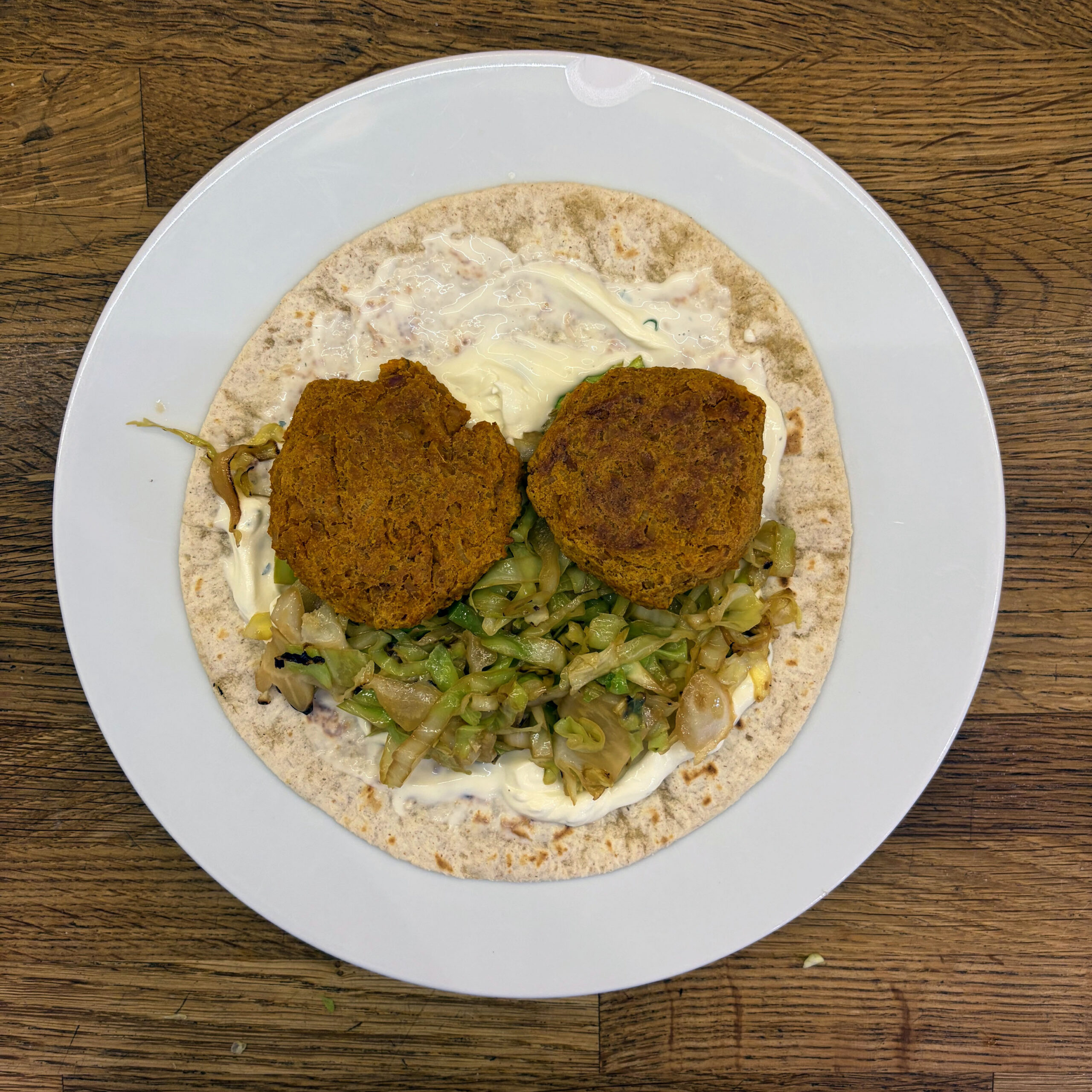 Zwei goldbraun gegrillte Kichererbsenpatties auf einem Weizenpitabrot, angerichtet mit sautiertem Weißkohl und einem cremigen Minze-Joghurt-Dip, serviert auf einem weißen Teller.