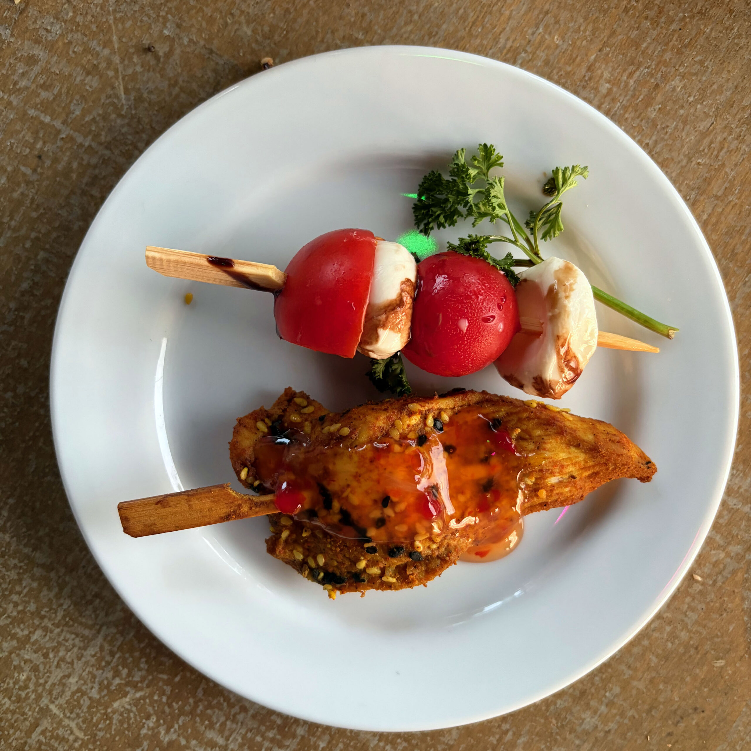 Weißer Teller mit zwei Fingerfood-Spießen vom Geburtstagsparty-Buffet: ein Tomaten-Mozzarella-Spieß mit frischer Petersilie und ein knuspriger Hähnchen- oder Fleischspieß mit Sesam und süß-scharfer Chilisauce.