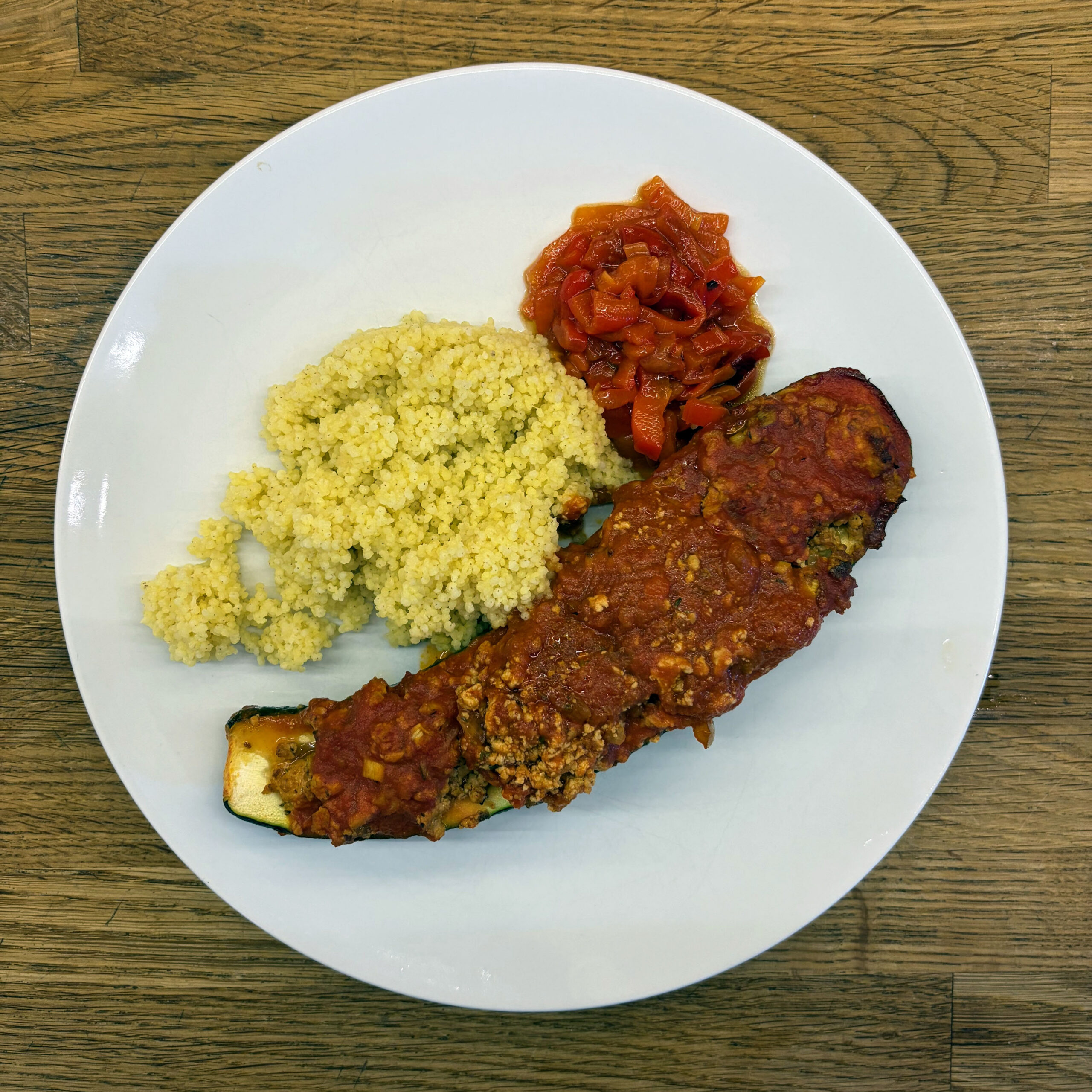 Weißer Teller mit einer mit Tofu-Tomaten-Füllung gefüllten und überbackenen Zucchinihälfte, serviert mit Hirse und einem Paprika-Tomatensalsa.