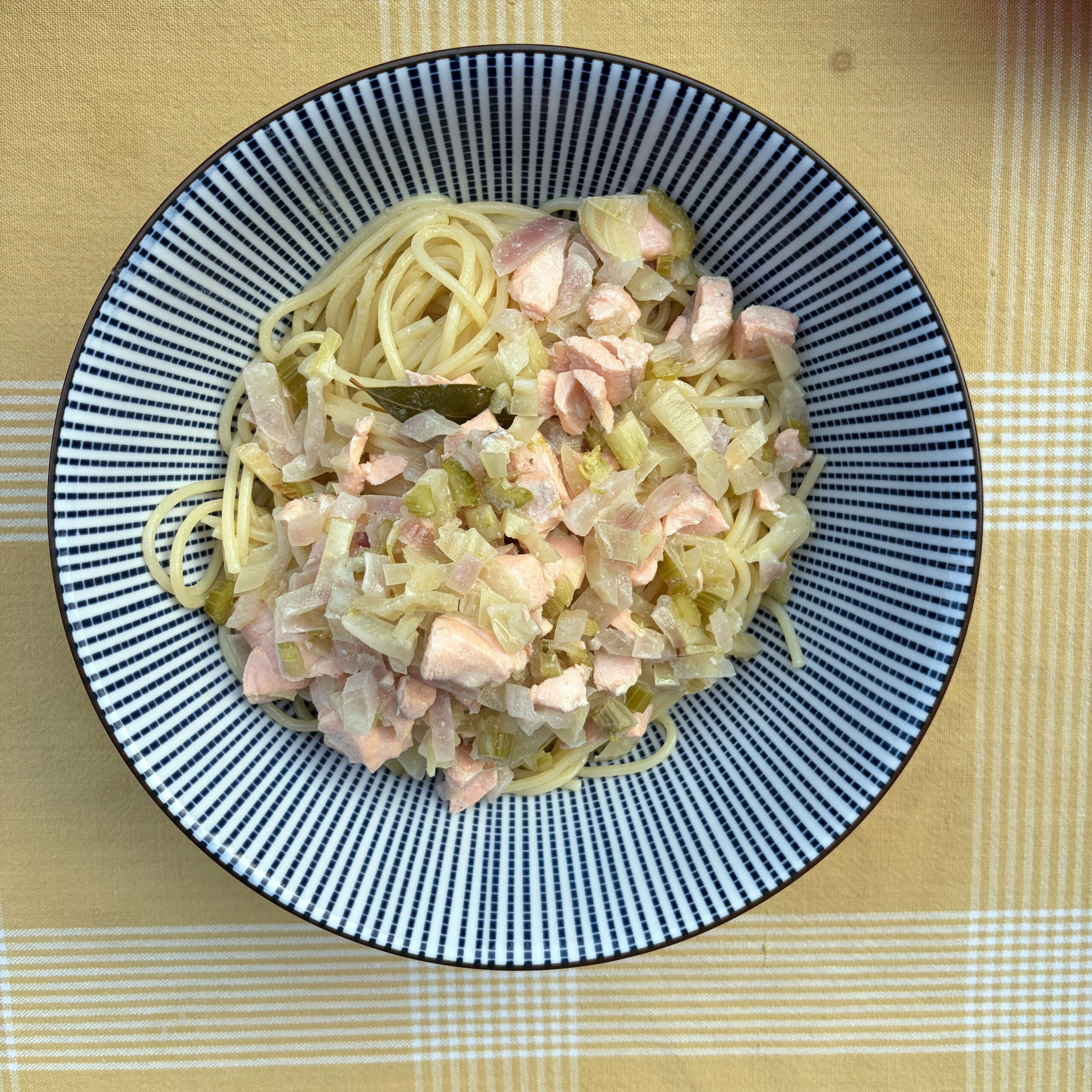 Draufsicht auf einen schwarz-weiß gestreiften Teller mit Spaghetti, belegt mit einer cremigen Fenchel-Lachs-Soße aus Lachsstücken, Fenchel, Sellerie und Zwiebeln, auf einem gelb-karierten Untergrund.