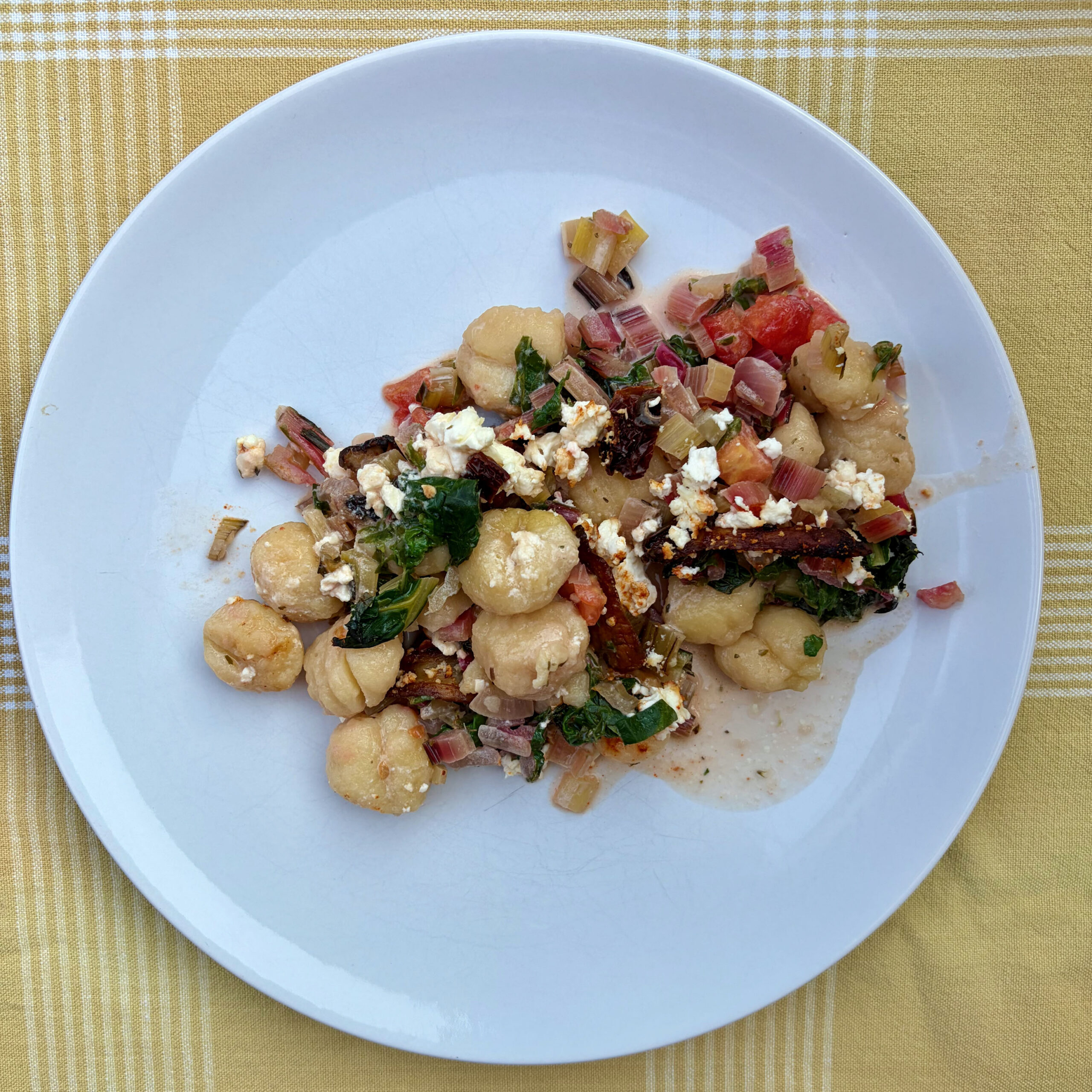 Weißer Teller mit gebackenen Gnocchi, sautiertem Mangold, Tomatenwürfeln, gebratenen Kräuterseitlingen und zerbröckeltem Feta auf gelbem Karostoff.